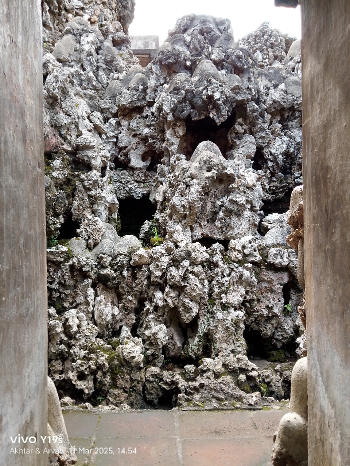 The image shows a stone wall made of rough, gray, and white rocks, with some small, dark crevices and green moss patches. The wall is viewed through a narrow opening, framed by two vertical concrete walls. The ground in front of the wall is made of reddish-brown tiles, with a white line running across it. The location, based on the coordinates provided, is likely somewhere in the city of Cirebon, Indonesia. There are no people or animals in the image.