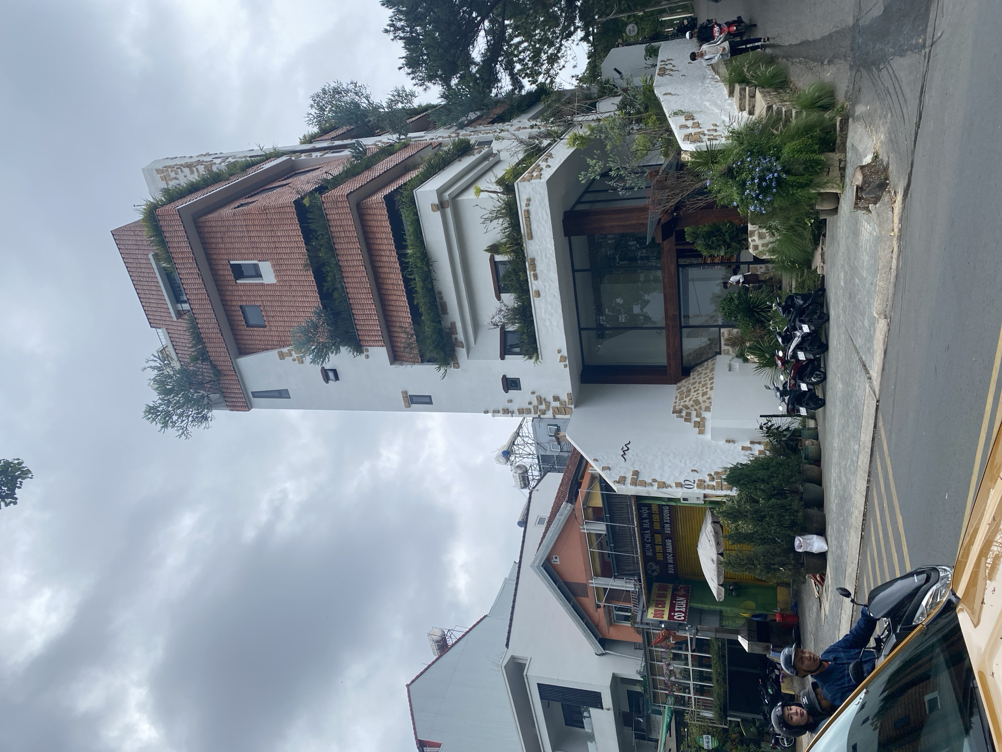 Here's a detailed description of the image:
**Setting:**
The image is taken on a street in Đà Lạt, Lâm Viên, Vietnam. The weather appears cloudy and overcast.
**Subjects and Objects:**
* **Unique Building:** A distinctive, multi-story building dominates the frame. It has a white exterior and terraced roof sections covered in red tiles. Each terrace also features greenery, creating a "living" architecture design.
* **Motorbikes:** Several motorbikes are parked near the building, indicating street-side parking common in Vietnam.
* **Adjacent Buildings:** A glimpse of other buildings are visible. One is colorful with commercial signage.
* **People:** In the foreground, a few people are on a motorbike and other individuals are walking on the sidewalk.
* **Street:** The image captures a segment of the street with parking lines and a partial view of a vehicle.
* **Vegetation:** Besides the greenery on the building, there are other plants, bushes, and trees surrounding the building and along the street.
* **Commercial Signage:** A building in the foreground displays Vietnamese text that includes "Bún Chả Hà Nội", referring to a popular Vietnamese dish.
**Composition:**
The photograph is taken from street level and depicts a mix of urban life and unique architecture. The cloudy sky serves as a backdrop. The street and parked vehicles provide foreground elements.