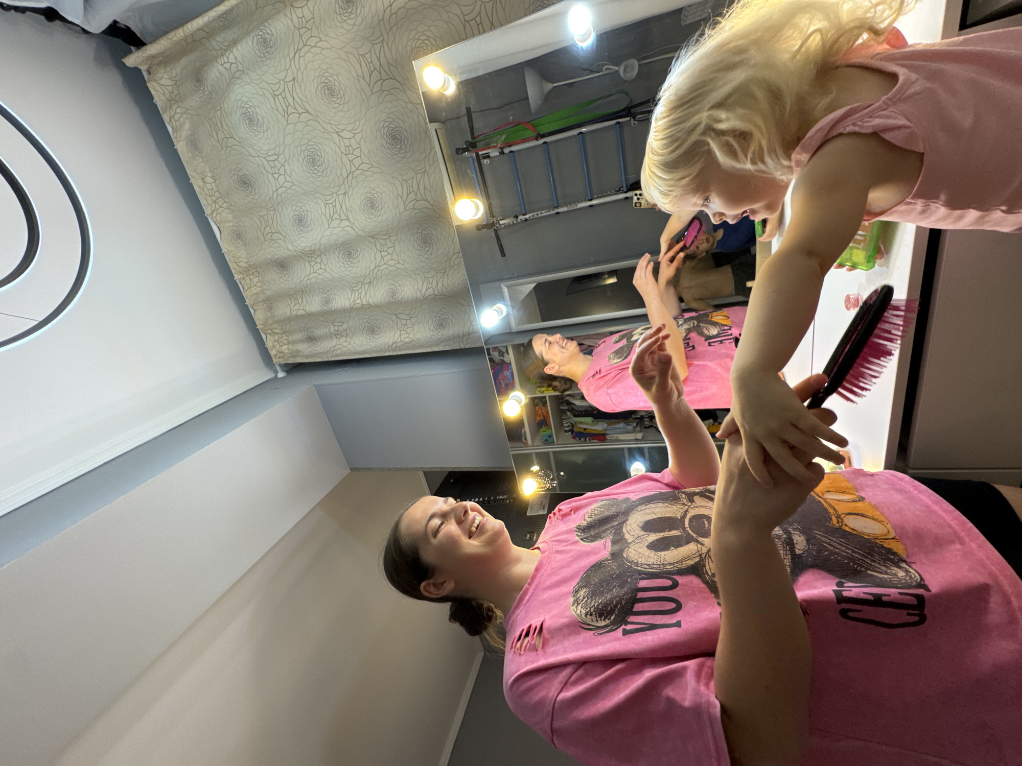 The photograph captures a woman in a pink t-shirt with a graphic of Mickey Mouse and a child with blonde hair, also in a pink top, interacting in front of a large mirror surrounded by round lights. The woman is smiling and looking up at the child, who appears to be brushing her hair with a black and red brush. The mirror's reflection shows a wider view of the room, including a curtain and a ladder-like structure on the wall. The lighting is bright, suggesting an interior setting. The city of Sochi, Russia is not recognizable in this image.