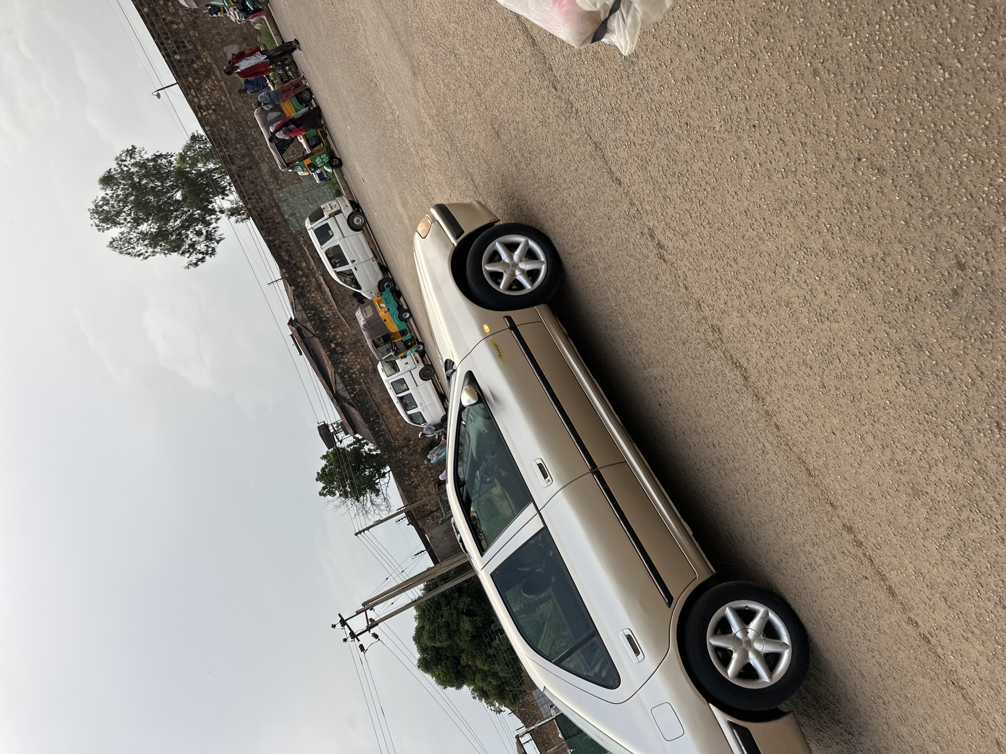 In Bukuru, Nigeria, a gold sedan is parked on a dusty, asphalt road. The car, which appears to be a Peugeot 406, is positioned diagonally across the frame, with its front facing to the right. Several auto-rickshaws, commonly known as Keke Napeps, are visible in the background, some carrying passengers. A few individuals are standing near the vehicles, including a man in a red jacket and another in a white shirt and trousers. In the distance, a low, stone wall runs horizontally, with a large tree and buildings behind it. Overhead, a cloudy sky dominates the upper portion of the image, with utility poles and power lines visible on the left side. The overall scene depicts a typical street setting in an urban area of Nigeria, likely a transportation hub or market area.