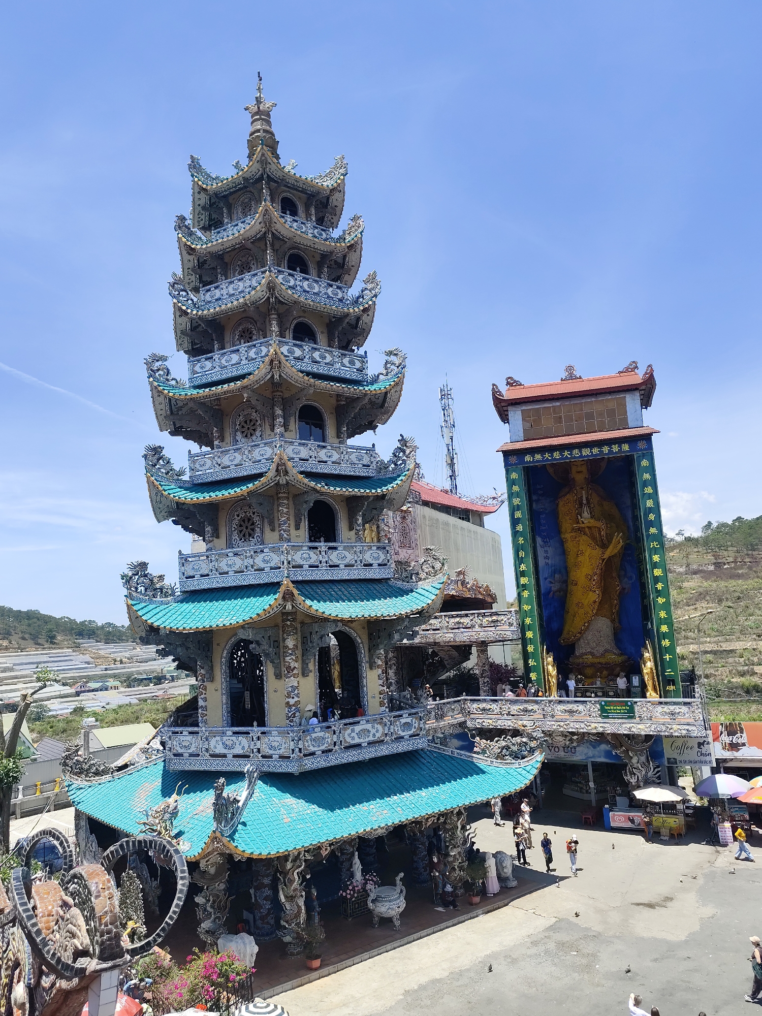 The image shows a tall, ornate pagoda and a large statue of a deity, possibly Buddha, at a location in Xuân Thành, Vietnam. The pagoda features multiple tiers with detailed decorations in blue, green, and beige, while the statue is housed in a structure with Chinese characters written on the front. People can be seen walking around the base of the structures, and there are also several businesses and shops, including a coffee shop. The overall environment suggests it is a tourist attraction, with a combination of religious structures and commercial activity.