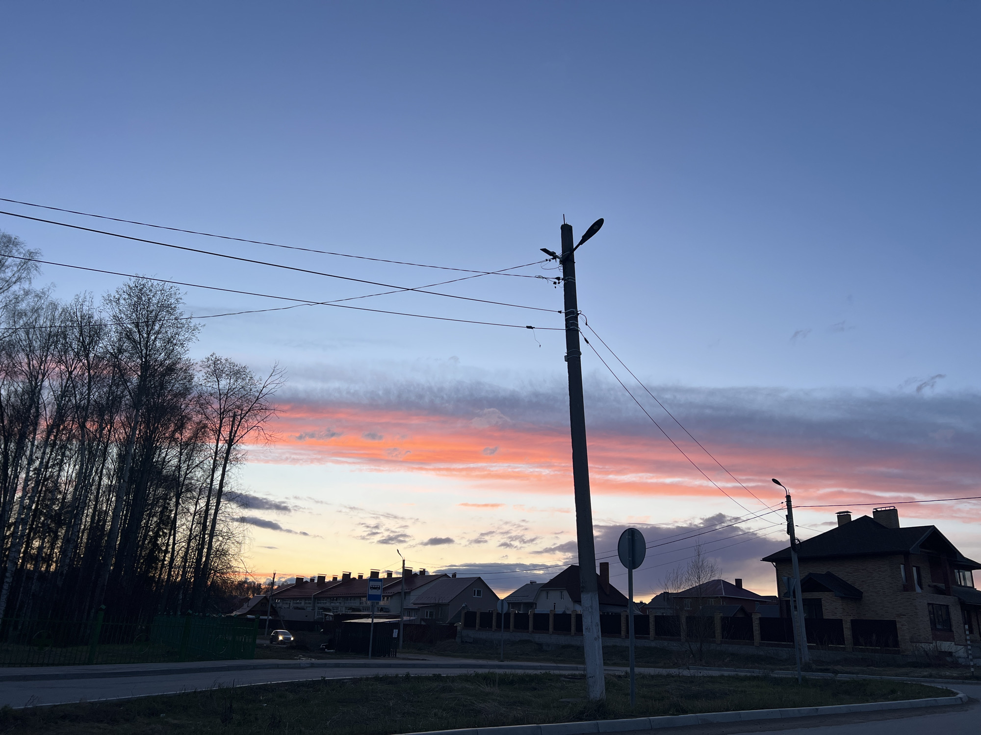 The image displays a serene sunset over a residential area in Noviy, Russia. The sky transitions dramatically from a deep indigo blue at the top to vibrant bands of orange, pink, and yellow closer to the horizon, with several elongated clouds streaking across the colorful expanse.
In the foreground, a prominent utility pole, equipped with multiple streetlights and numerous power lines, stands slightly right of the frame's center, creating a dark silhouette against the bright sky. To the left, a dense group of tall, bare-branched trees, suggesting late autumn or early spring, also forms a dark mass. A green metal fence runs along the bottom edge of these trees, adjacent to a paved road and curb.
Further into the scene, several residential houses are visible, featuring dark roofs and mostly two-story structures. A large brick-faced house with multiple chimneys is prominent on the far right. Another utility pole and a circular road sign, whose text is unreadable, are also present. In the mid-distance on the left, a bus stop shelter can be seen. A single car with its headlights illuminated is visible on the road in the distance, indicating its movement in the dimming light. The streetlights on the utility poles are not lit, suggesting that natural light from the sunset is still adequate.
The scene depicts the quiet transition from day to night in a suburban or semi-rural setting, characterized by the striking colors of the sunset, the bare trees, and the architectural styles of the houses. No people are visible.
