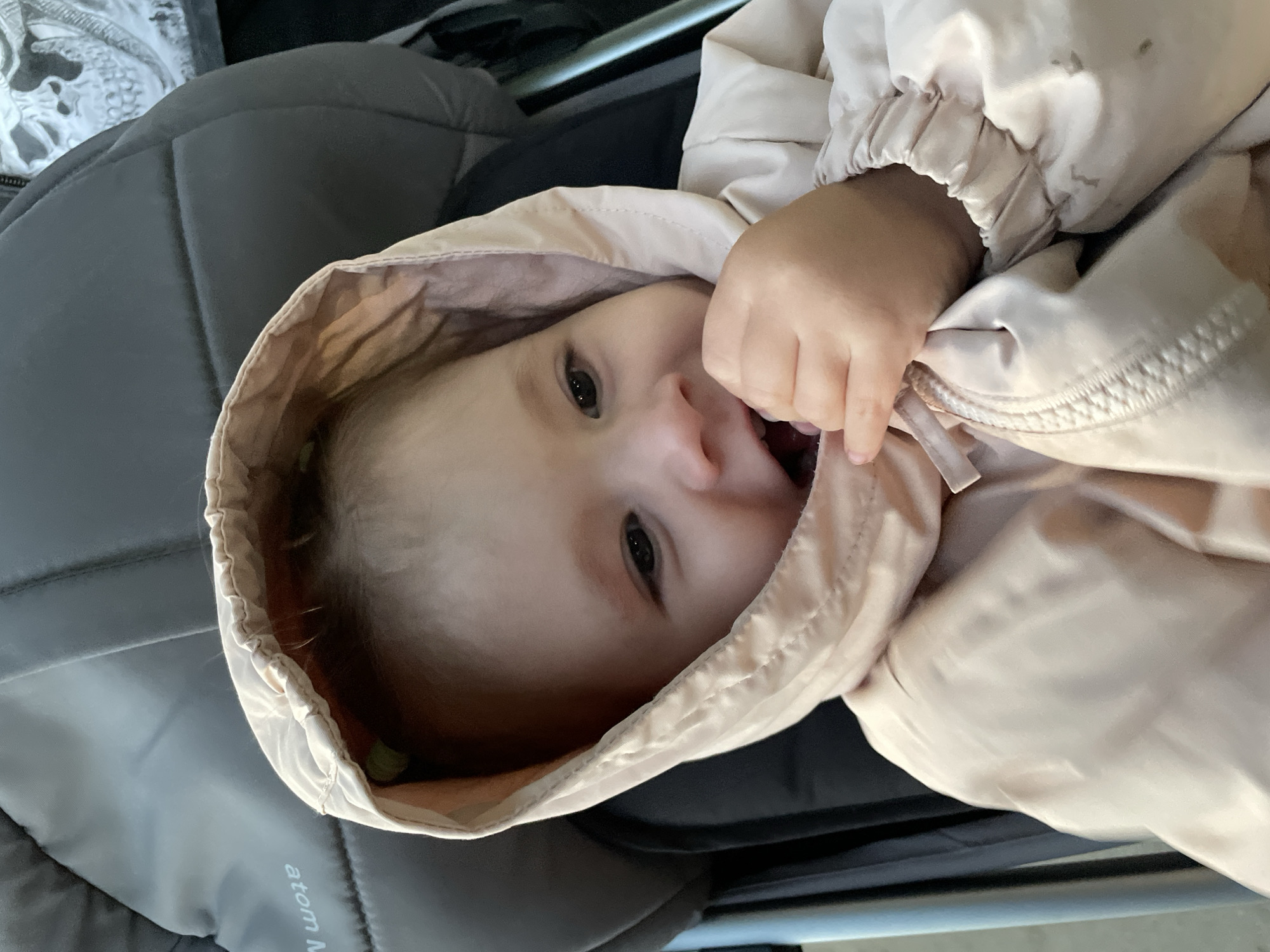 A young child, possibly an infant or toddler, is lying down and partially obscured by a light pink or beige hooded jacket. The child's face is visible, displaying a curious expression with eyes open and mouth slightly agape. One hand is raised, holding onto the fabric of the jacket. The child is positioned in what appears to be a gray padded surface, possibly a car seat or stroller. Metallic bars are visible in the background, suggesting a frame or support structure. The exact location within Izhevsk, Russia, is not discernible from the image alone.