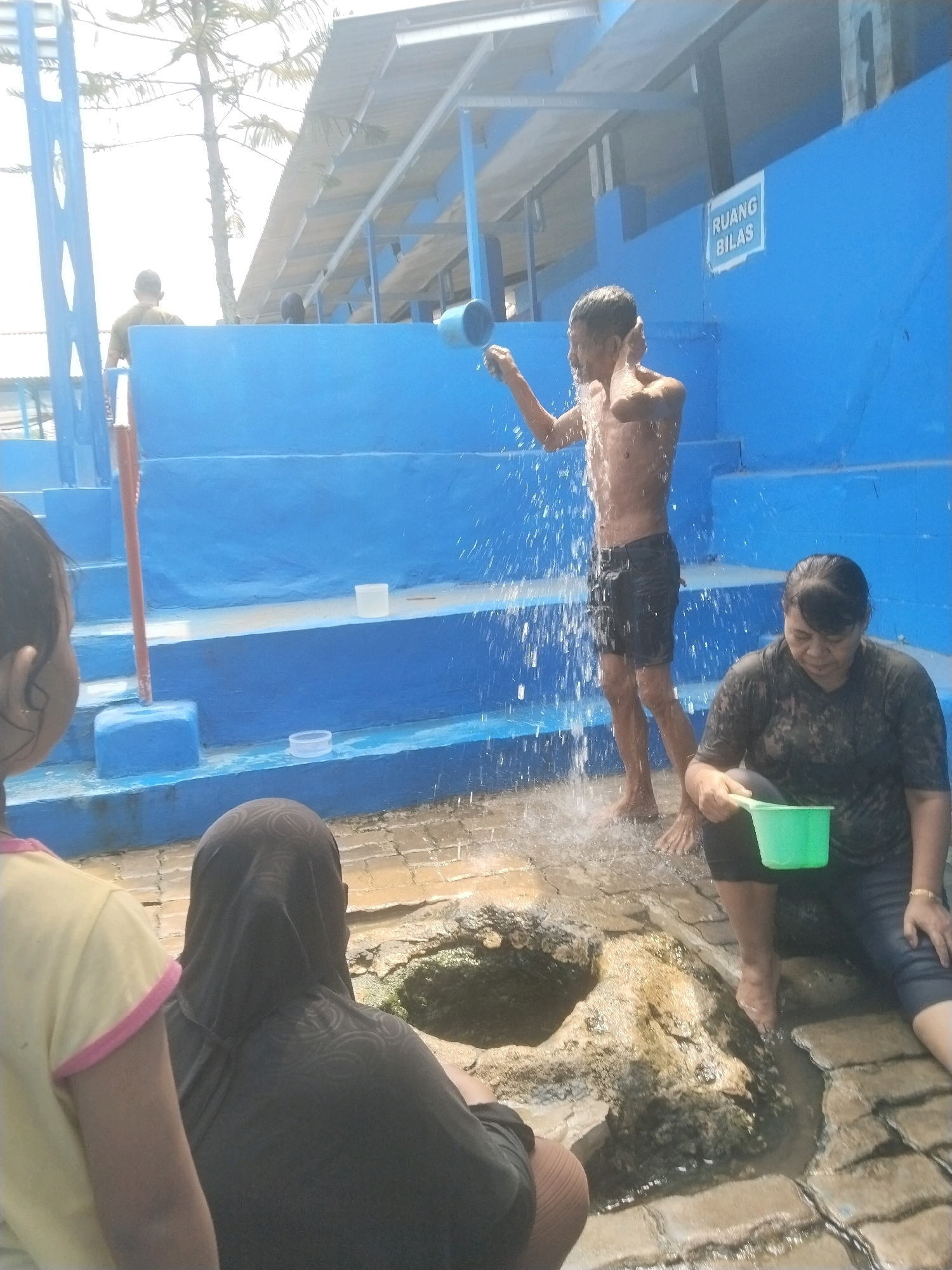 The image depicts an outdoor scene at what appears to be a public bath or water park facility in Batukuwung, Indonesia. In the background, vibrant blue tiered structures suggest swimming pools or changing areas. A young man, shirtless and wearing dark shorts, is actively showering himself with water from a blue bucket, creating a spray of water around him. To the right, a woman, also wet, is seated on the ground, holding a green plastic bucket. In the foreground, two individuals are partially visible. One, on the left, is a child wearing a yellow shirt with pink trim, facing away from the camera. The other, in the center, is a person wearing a dark hijab and headscarf, also facing away. The ground is paved with interlocking bricks, and a naturalistic rock formation with water flowing around it is present in the lower center of the frame. The overall atmosphere suggests a recreational or communal bathing area.