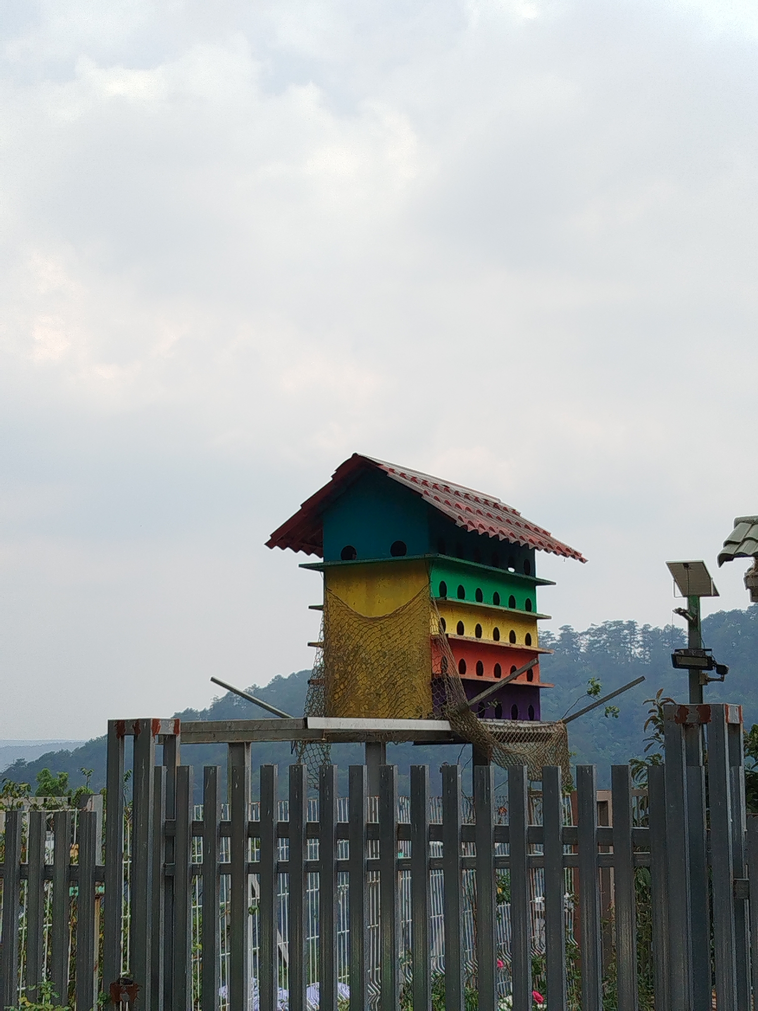 The image captures a multi-colored, multi-tiered birdhouse, situated behind a metal fence. The birdhouse consists of several levels, each painted a different color (teal, green, yellow, orange, red, and purple), and topped with a red-tiled roof. A net-like structure hangs down one side of the birdhouse. The fence is made of dark vertical bars. In the background are distant mountains and a cloudy sky. A metallic object with a solar panel is visible to the right of the birdhouse. This scene is identified as being in Da Lat, Vietnam.