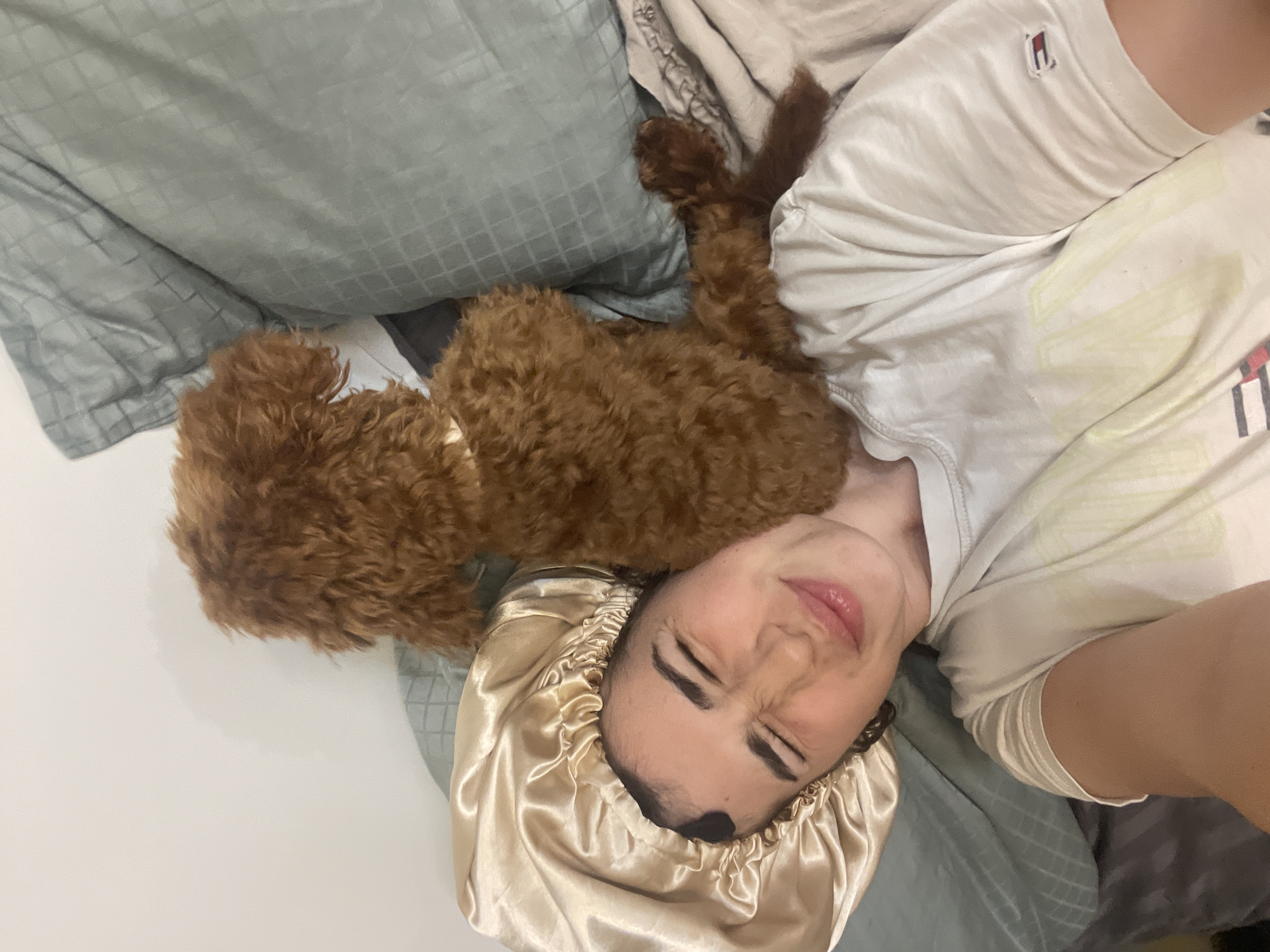 This indoor image, set in what is likely a home environment in Sofia, Bulgaria, captures a person and their dog relaxing on a bed or sofa. The perspective is an overhead shot, looking down at the subjects.
The main subject is a person, likely female, lying horizontally, wearing a light-colored, short-sleeved t-shirt. The t-shirt features a small, distinctive rectangular logo on the upper chest, displaying red, white, and blue stripes, which is identifiable as the Tommy Hilfiger brand. Her head is resting on a pillow, covered by a shiny, light gold or champagne-colored satin sleep bonnet. Her eyes are closed, and her facial expression shows a slight grimace or pucker of the lips, which could indicate sleepiness, a moment of discomfort, or a playful contortion.
Snuggled closely to the person's head, on the same pillow, is a small to medium-sized dog with dense, curly, reddish-brown fur, consistent with a poodle or a doodle breed. The dog is lying down, appearing relaxed next to its companion.
The setting is a cozy bedroom or living area, with crumpled pillows and bedding in muted tones, including a light blue pillow with a subtle checkered pattern and off-white sheets. The lighting is soft and indirect, suggesting an indoor ambiance, possibly morning or evening. The overall scene conveys a sense of comfort and companionship between the person and their pet. No other specific location details are visible within the frame.
