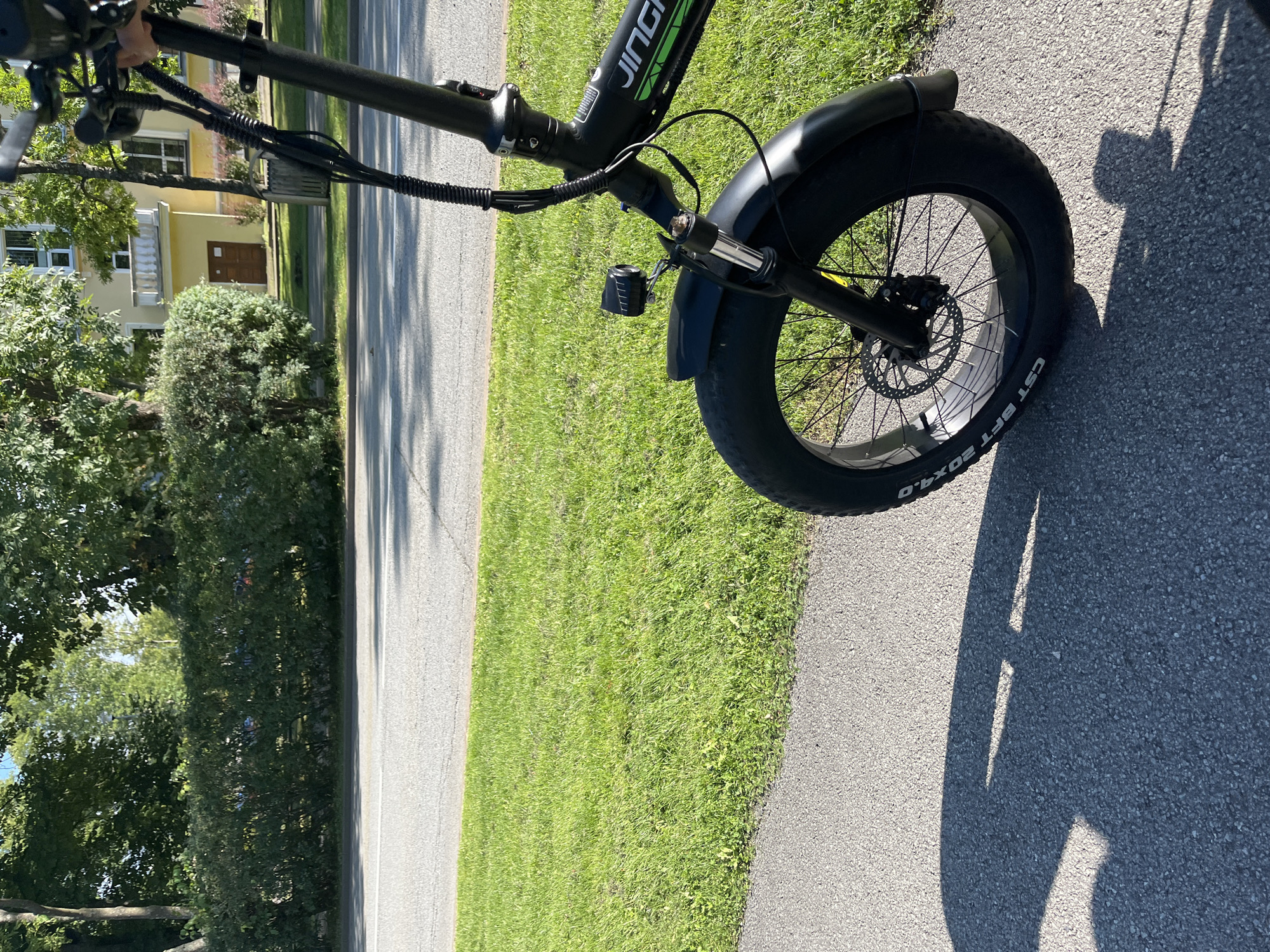 The image depicts a black electric bicycle parked on a paved pathway in the city of Kohtla-Järve, Estonia. The bicycle is the primary subject, with its front wheel, fork, handlebars, and part of the frame visible. A small black headlamp is attached to the front fork. The bicycle has a prominent fat tire.
Behind the bicycle, a well-maintained green lawn stretches to a road. Across the road, a hedge forms a dense green barrier, beyond which a light yellow building with white trim and windows can be seen. Trees with lush green foliage are scattered throughout the background, suggesting a warm, sunny day. The shadow of the bicycle, along with what appears to be a person's shadow holding the handlebars, is cast on the pathway in the foreground. The overall scene is peaceful and urban, with elements of both nature and man-made structures.