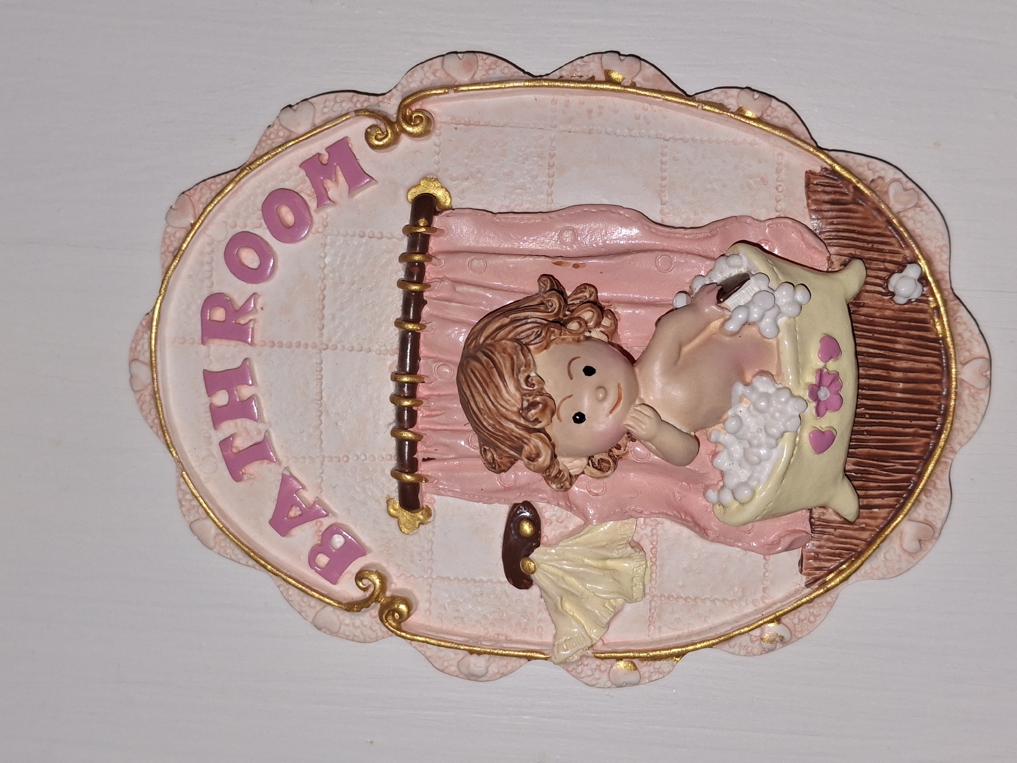 This is a decorative plaque that says "BATHROOM" in pink lettering. The plaque features a pink background with a textured pattern and a gold border. In the center, a small figurine of a baby is sitting in a bathtub filled with bubbles. The baby is holding a brush. A pink curtain hangs behind the bathtub. A cream-colored towel is draped over a hook to the left of the curtain. The plaque is oval-shaped with a scalloped edge. The plaque is mounted on a white wall. The location in Åsgårdstrand, Norway is not visible in the image.