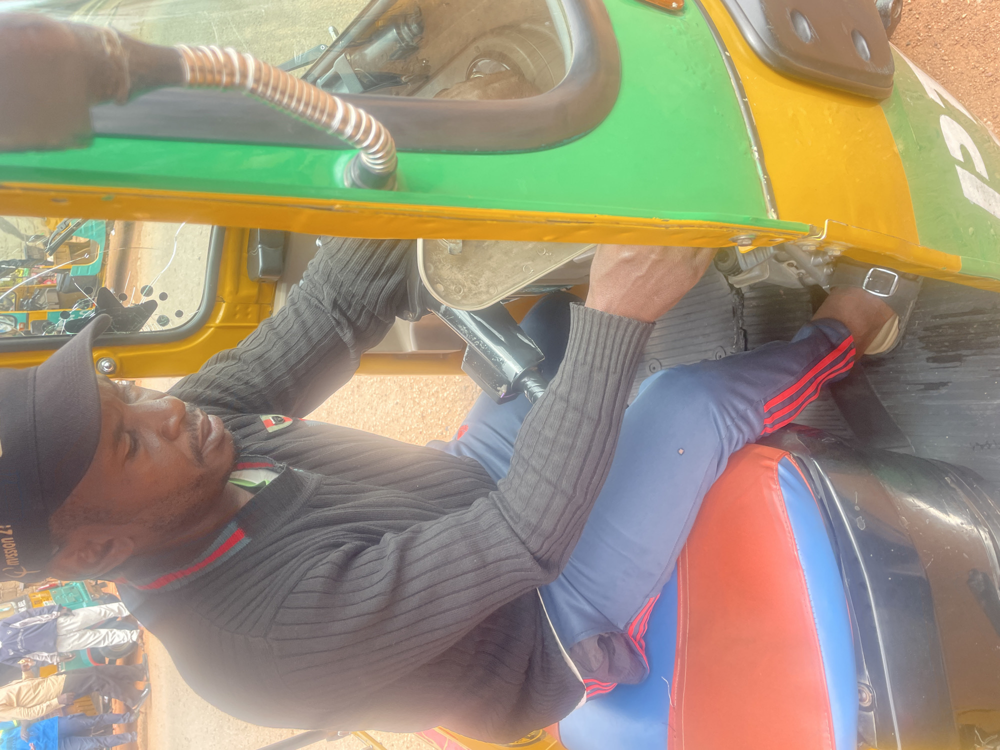 In Bar Jirgi Summa, Nigeria, a man wearing a black knit sweater and blue track pants with red stripes sits in the driver's seat of a yellow and green auto-rickshaw. He is looking down and appears to be operating the vehicle's controls with his right hand. The auto-rickshaw has a cracked windshield and a colorful blue, red, and orange seat. In the background, several other people and auto-rickshaws are visible, suggesting a busy street or market scene. The ground is covered in reddish-brown dirt.