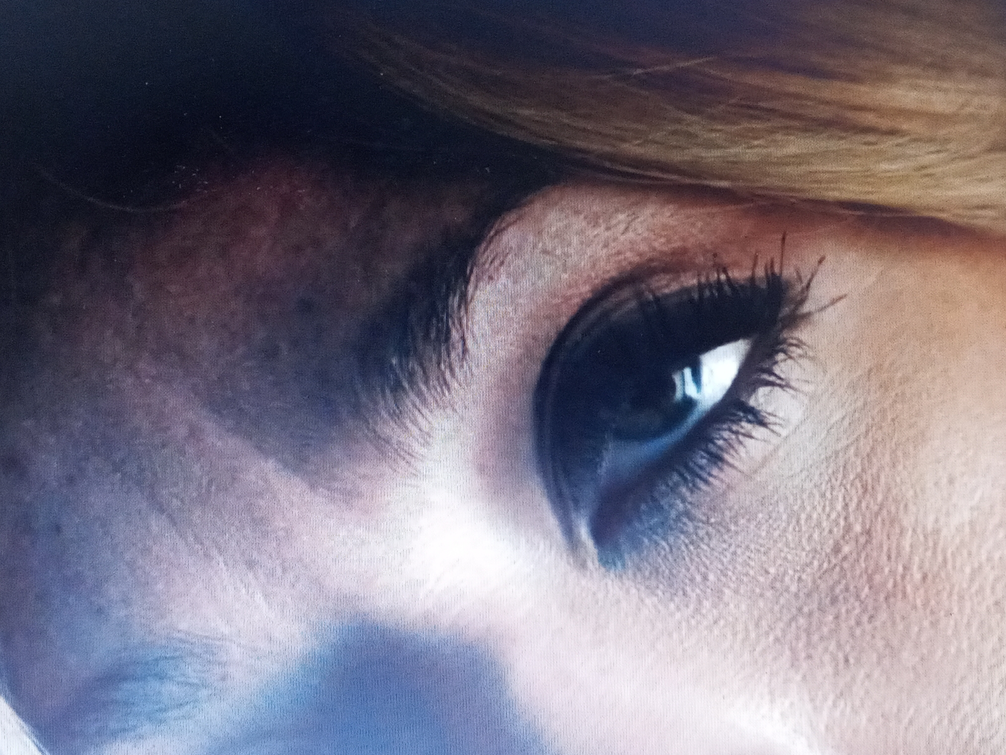The image shows a close-up of a person's eye and surrounding facial features. The eye is dark, with visible eyelashes and a reflection in the pupil. The skin around the eye is textured, with visible pores and fine lines. Part of an eyebrow is visible above the eye, and some hair is seen at the top of the frame. The overall lighting is soft, with shadows and highlights accentuating the contours of the face. No location is discernible from the image.