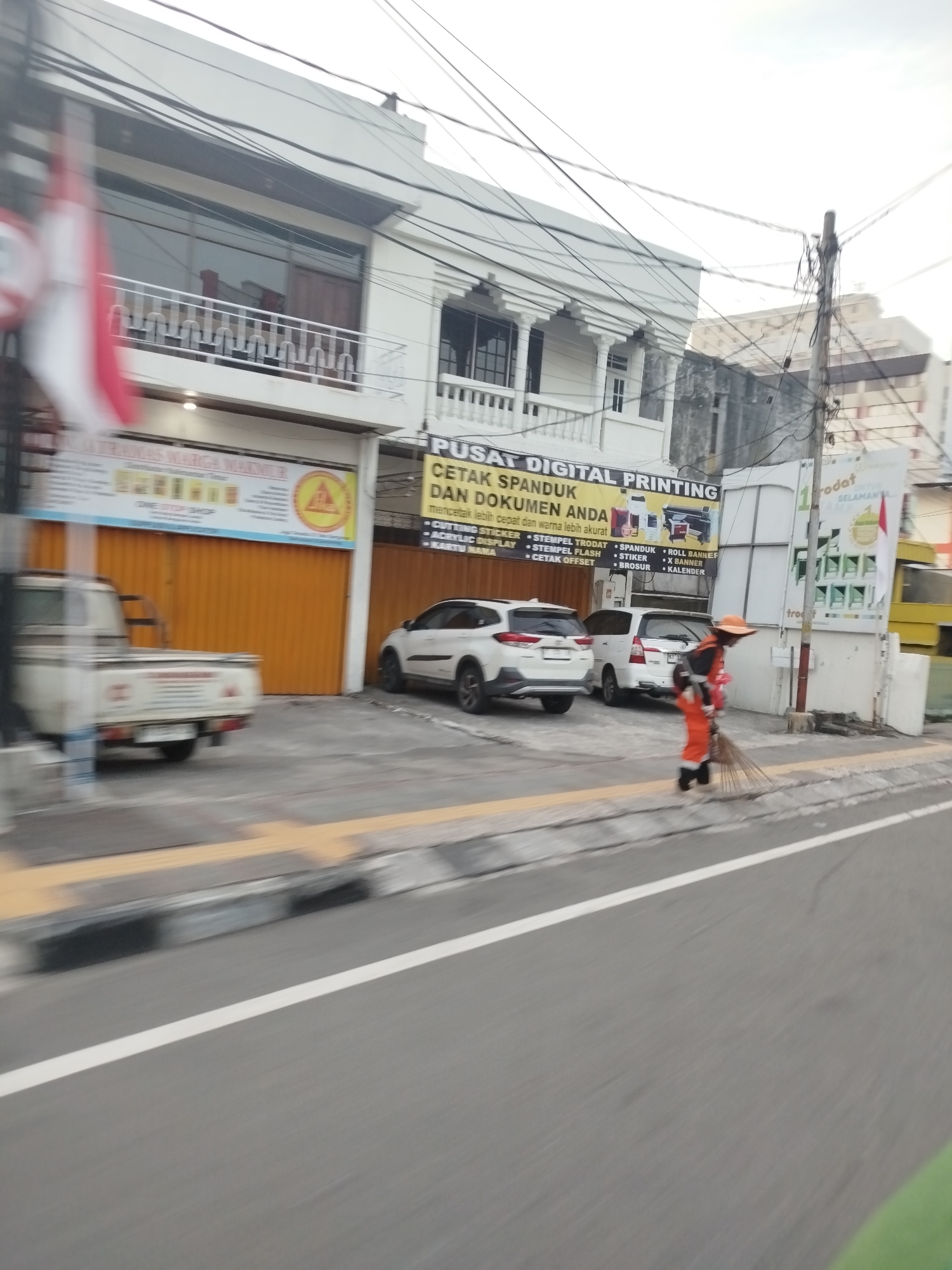The image shows a street scene in Balikpapan, Indonesia, with a blur of motion suggesting it was taken from a moving vehicle. In the foreground, the asphalt road is visible with a white lane marker. A sidewalk runs along the right side of the road, with a curb and a yellow tactile paving strip. Behind the sidewalk, there are several buildings.
One building on the left has an orange-colored corrugated metal garage door and a sign that reads "TRANS MARGA MAKMUR" with a yellow circular logo. Above this, a white facade with a balcony and railing is visible.
To the right of this, a building with a white facade has a sign that reads "PUSAT DIGITAL PRINTING CETAK SPANDUK DAN DOKUMEN ANDA" followed by smaller text and images of printing equipment. This building also has a balcony with columns and a railing.
Further to the right, a utility pole with a dense tangle of electrical wires stands near another building. A sign on this building, partially obscured by wires, includes the word "trodat" and an Indonesian flag. A yellow vehicle, possibly a bus, is parked behind this building.
In front of the "PUSAT DIGITAL PRINTING" building, two white cars are parked: a white SUV and a white minivan. A person in an orange uniform and hat is sweeping the sidewalk with a broom. The person appears to be wearing dark pants and white shoes. A small child, also wearing an orange outfit, is standing next to the person with the broom. The general environment suggests a commercial or mixed-use area in an urban setting.