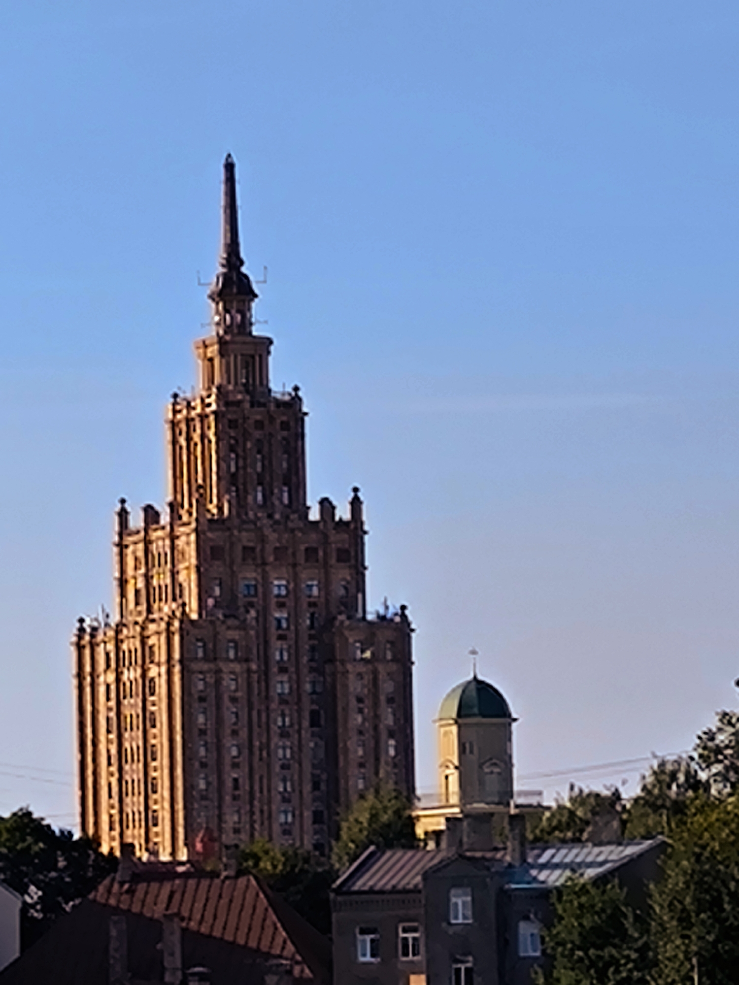 This image captures a view of Riga, Latvia, showcasing the Latvian Academy of Sciences building, a prominent Stalinist Empire style skyscraper, and a smaller, dome-topped structure to its right. The sky is a clear, pale blue, with the sun casting a warm, golden light on the facade of the main building, highlighting its architectural details and numerous windows. The skyscraper is centrally located and dominates the upper portion of the frame. To its right, a building with a distinctive green dome and a cross on top is visible, appearing shorter and less ornate than the main tower. In the foreground, the roofs of other buildings, some with darker, weathered surfaces and others with lighter, more modern materials, are visible. Trees with lush green foliage frame the lower part of the scene, suggesting a temperate climate and a time of year with abundant vegetation. The overall impression is one of urban architecture set against a clear sky, with the warm sunlight creating a sense of depth and highlighting the textures of the buildings. There are no people or animals visible in the image.