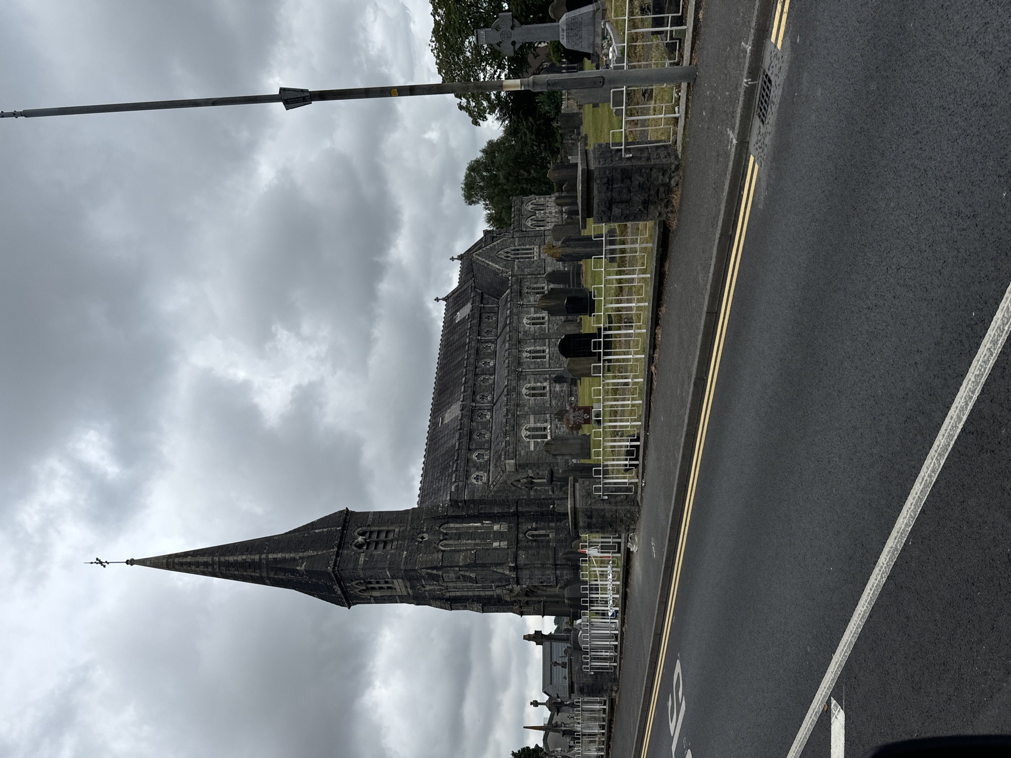 The image depicts a view of St. John's Cathedral in the city of Waterford, Ireland, from a road. The cathedral, constructed of dark stone, features a prominent steeple topped with a cross. Adjacent to the cathedral is a cemetery with numerous gravestones and a large Celtic cross. A white metal fence separates the cemetery from the road. The road itself has double yellow lines painted on it, indicating parking restrictions, and a white line marking a lane. A street lamp stands on the right side of the image. The sky is overcast with heavy clouds. There are no people visible in the scene.
