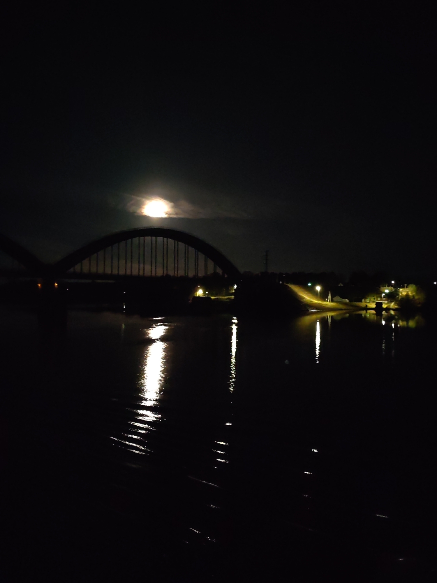 At night in Gladyshevo, Russia, a body of water reflects the light of a full moon obscured by clouds. A bridge spans the water, with its arched structure and vertical supports silhouetted against the night sky. Streetlights illuminate a road and surrounding buildings on the far side of the water. The water's surface shows rippling reflections of the moon and the streetlights.