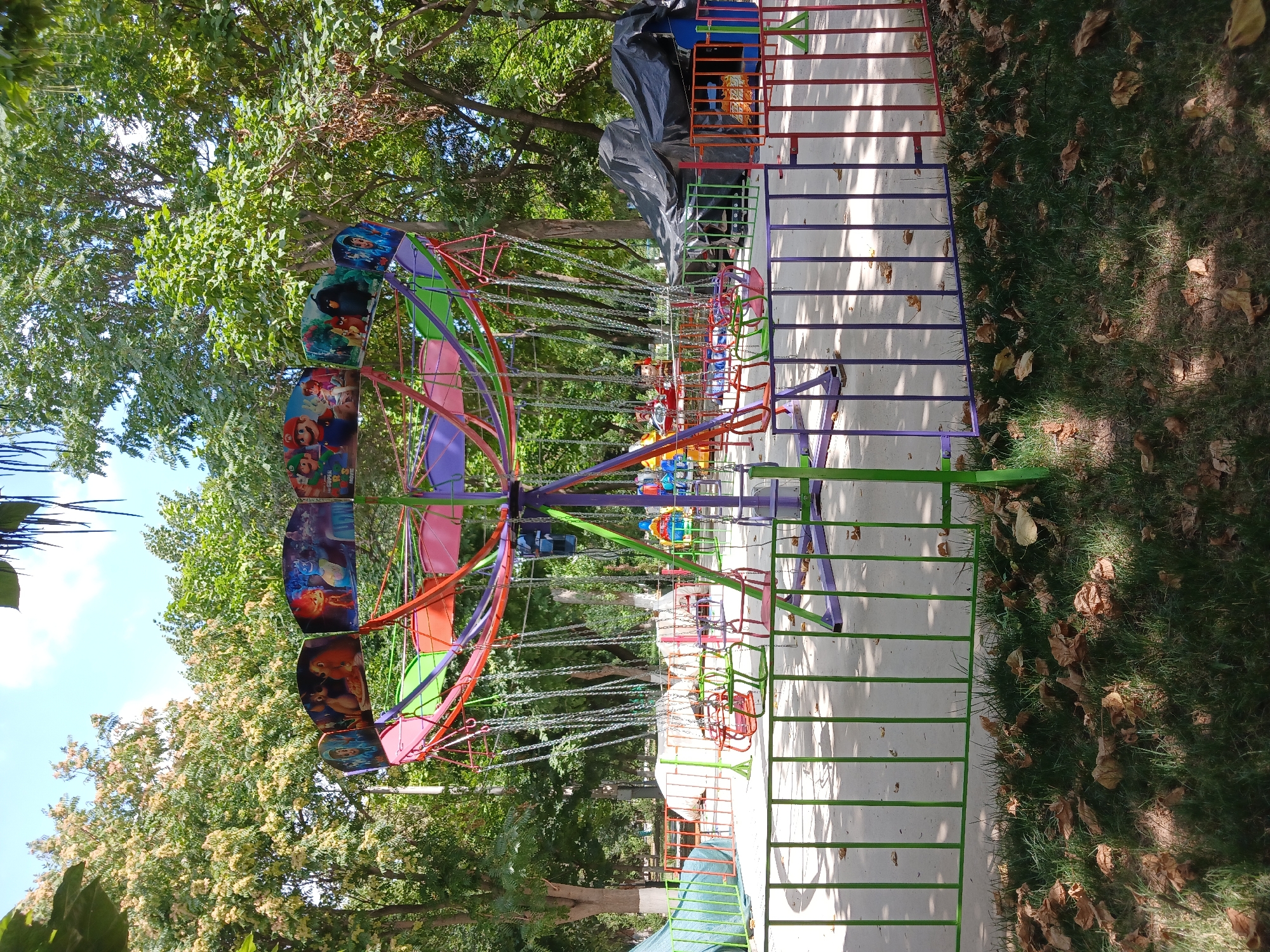 A colorful amusement park ride, likely a swing carousel, stands in an outdoor setting. The ride features a central pole with a rotating canopy decorated with cartoon characters, including Mario from Super Mario and characters from "The Secret Life of Pets" and "Lady and the Tramp." The swings themselves are red, green, and pink. Surrounding the ride is a fence with vertical bars in green, purple, and red. The ground is paved, and dry leaves are scattered on the grass in the foreground. Trees with lush green foliage are visible in the background, suggesting a park or recreational area. The sky is blue with some clouds. This scene is located in the Mirzo Ulugbek District of Tashkent, Uzbekistan, within the Tashkent Land of an Amusement park. There are no people visible in the image.