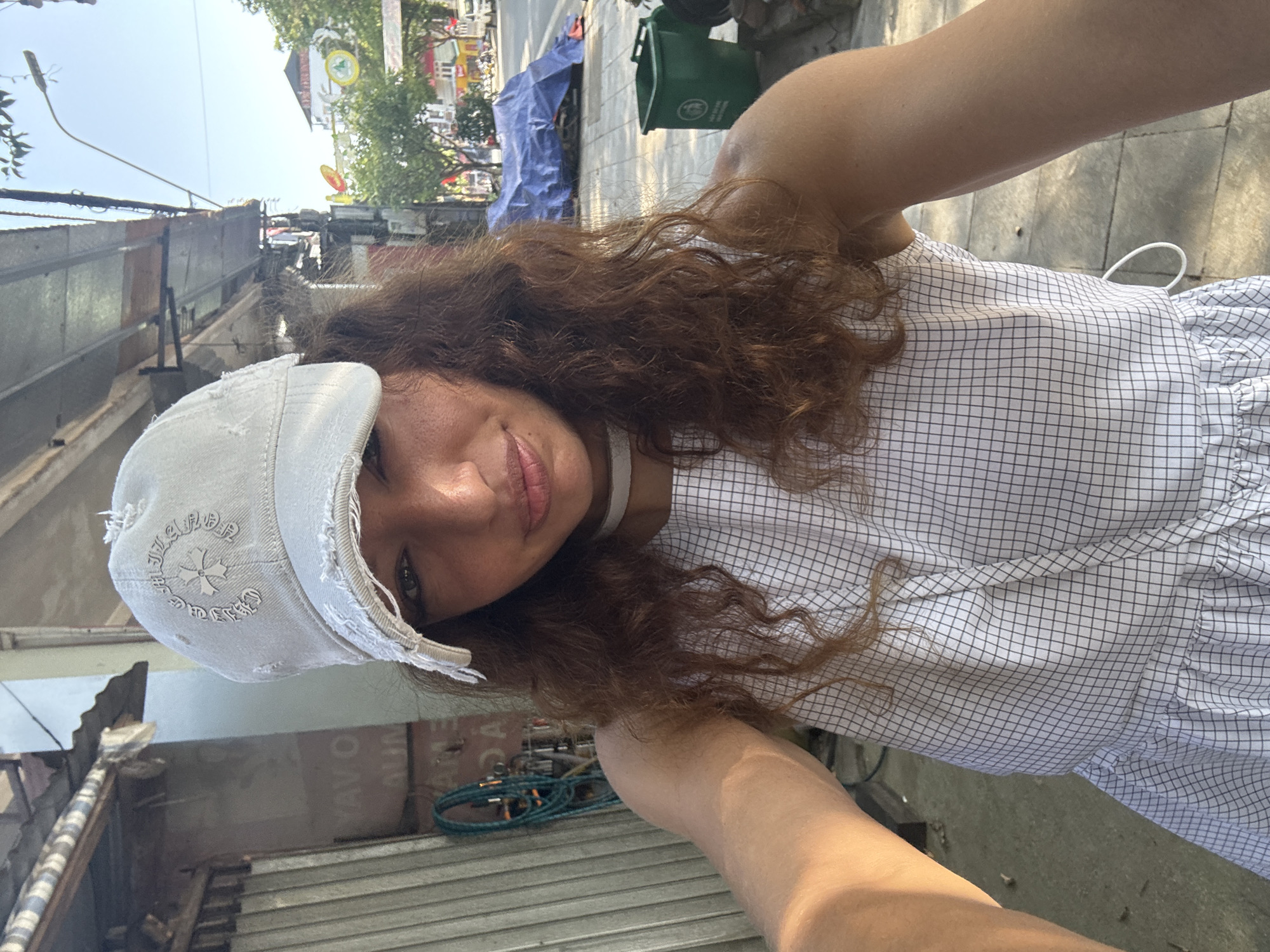 A woman with curly brown hair and fair skin wears a white cap and a white and black grid-patterned dress, likely taking a selfie in Bắc Hà, Vietnam. She is positioned outdoors on a sidewalk bordered by buildings of varying materials, including corrugated metal and a blue tarp-covered area. A green trash bin is visible behind her. The lighting suggests it is daytime.