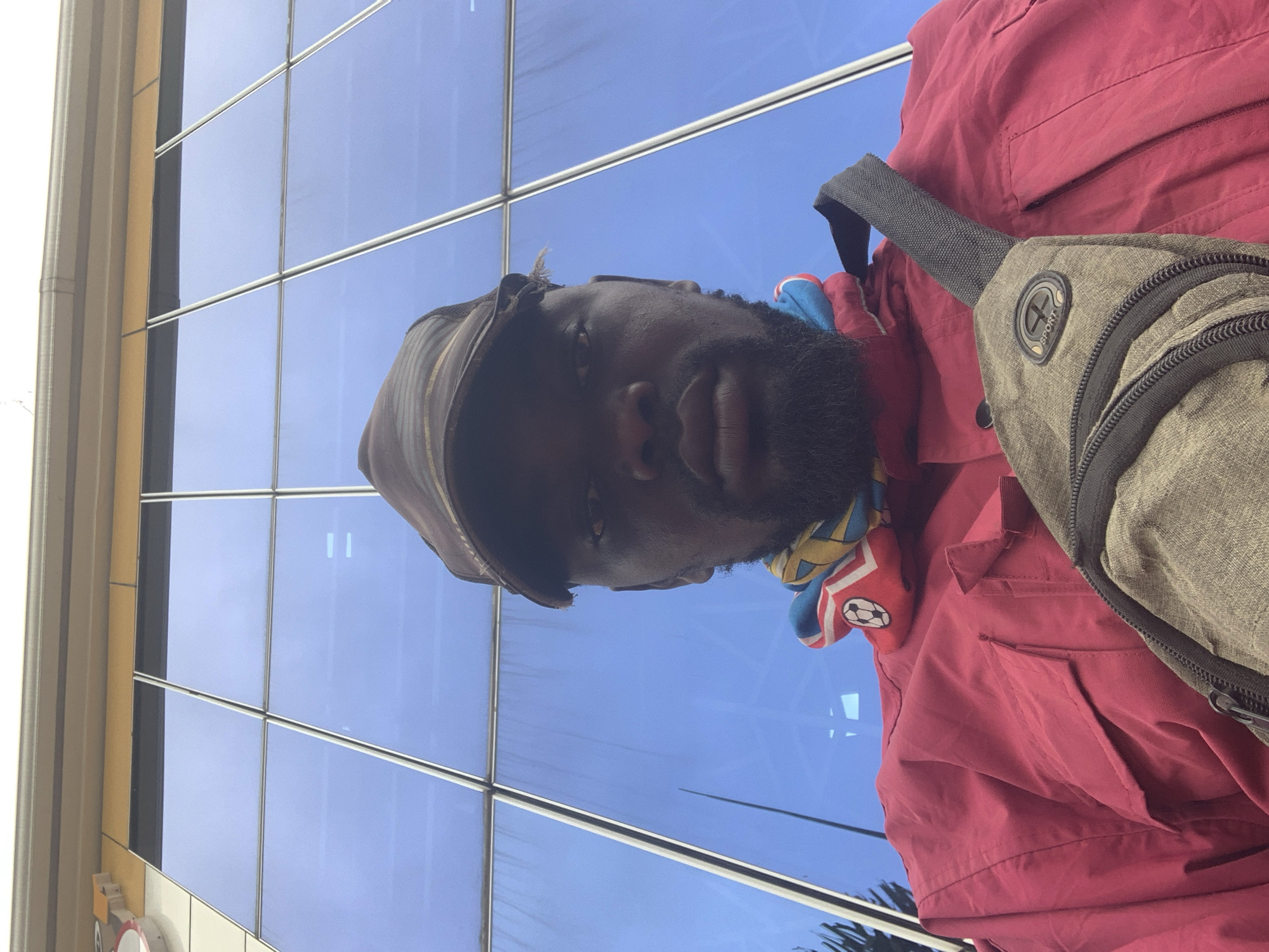 The image shows a low-angle, close-up, selfie-style shot of a Black man in Shagamu, Nigeria. He is wearing a camouflage-patterned baseball cap, a red jacket, and a grey cross-body bag. A colorful scarf with a soccer ball emblem is visible around his neck. He has a beard and is looking directly at the camera. The background consists of large, reflective blue glass windows of a building, suggesting an urban setting. The building appears modern, with a clean facade. The lighting is overcast.
