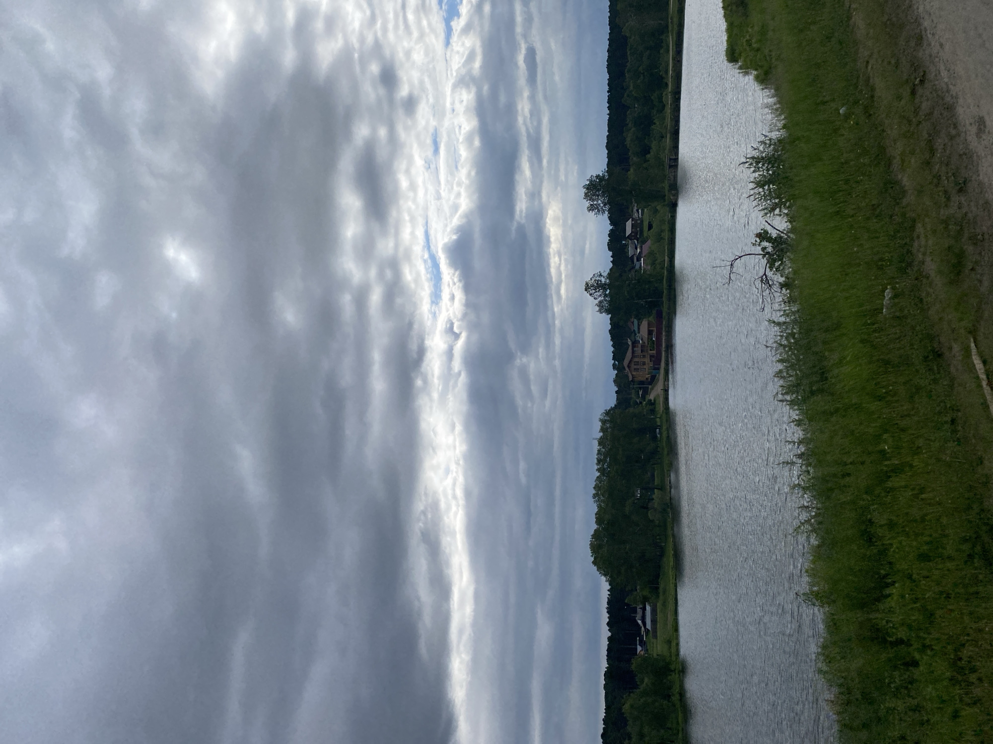 The image depicts a body of water, likely a lake or river, with a shoreline covered in green grass and vegetation. In the background, across the water, is a small settlement with houses nestled amongst trees. A dark, dense forest forms the horizon line behind the settlement. The sky is overcast with heavy, grey clouds, with some lighter patches suggesting the possibility of the sun breaking through. The overall impression is of a rural, natural landscape under a cloudy sky. The location is identified as Malta, Russia, suggesting a serene, possibly remote, Russian countryside setting. There are no people or animals visible in the image.