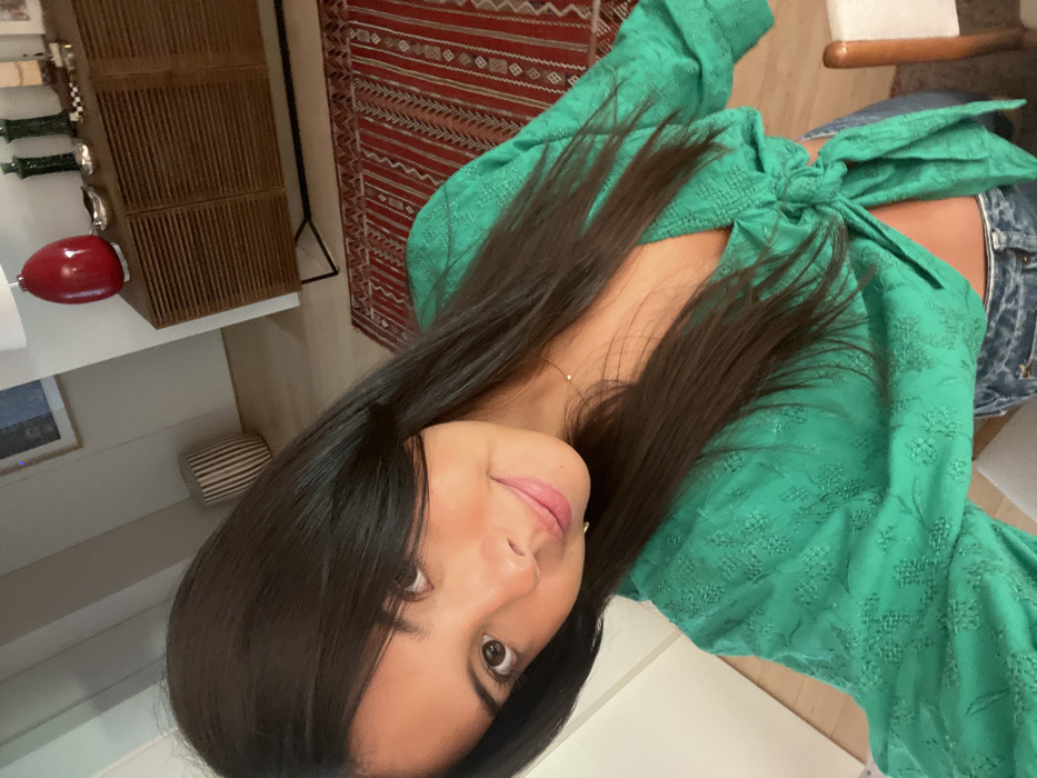 The image captures a young adult female in a selfie format, positioned in the foreground of an indoor residential setting. The specific location within Lordelo do Ouro e Massarelos, Portugal, cannot be determined from the visual information provided.
The subject has long, straight, dark brown hair, dark eyes, and a light-to-medium skin tone, exhibiting a subtle smile and direct eye contact with the camera. She is dressed in a textured, emerald green, long-sleeved, wrap-style crop top with a tie-front closure, revealing a portion of her midriff. Blue denim jeans are partially visible. A delicate gold-colored necklace with a small pendant is worn.
The background reveals a contemporary room featuring light-colored walls and light-toned wooden flooring. A red rectangular rug with intricate geometric patterns covers part of the floor. To the right, a wooden sideboard with slatted doors and black metal legs supports a red oval table lamp with a white shade, two tall dark green decorative vases, a silver-colored bowl, and a small checkered object. The upper left background shows a framed abstract artwork on the wall and a vertically striped cylindrical stool or decorative item. A section of a light-colored wooden chair with a white seat is visible in the bottom right corner. The environment is well-lit by ambient light.
