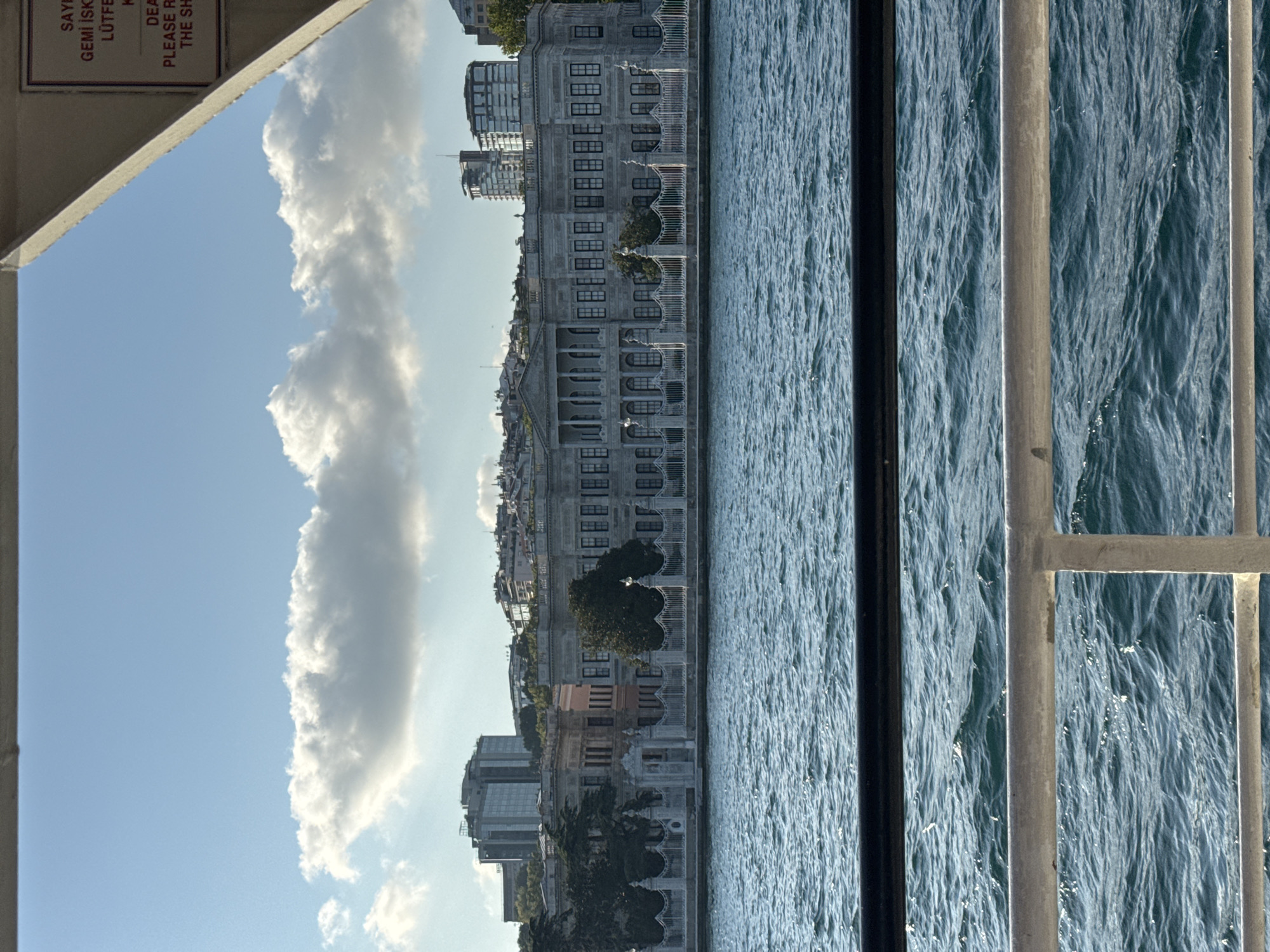 The image depicts a scenic view of the Bosphorus strait in Beşiktaş, Turkey, as seen from a ferry. In the foreground, a metal railing partially obstructs the view of the rippling blue water. Dominating the middle ground is the grand Dolmabahçe Palace, a magnificent 19th-century Ottoman imperial palace, characterized by its ornate architecture, classical columns, and numerous windows. The palace is set against a backdrop of lush trees and a white ornate fence. Beyond the palace, the cityscape of Beşiktaş unfolds with modern high-rise buildings and traditional rooftops. The sky above is a clear blue, punctuated by fluffy white clouds. A sign with text in Turkish and English is visible in the upper right corner of the image.