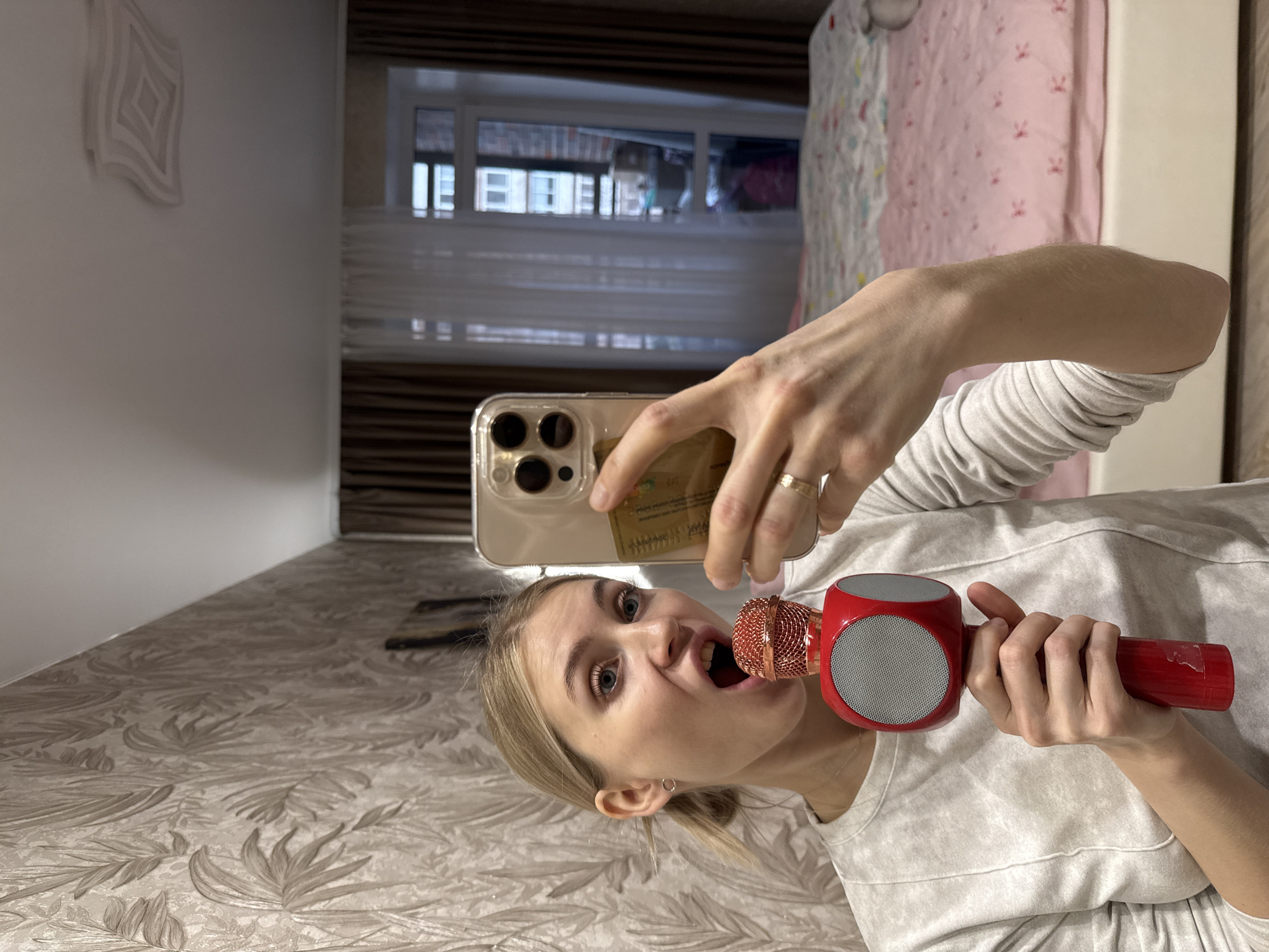 A young, fair-skinned woman with blond hair is holding a red microphone in her right hand and a smartphone in her left, seemingly taking a selfie. She is wearing a light-colored, long-sleeved shirt. Her mouth is open as if singing or speaking into the microphone. She has a ring on her ring finger. The smartphone case appears to be clear and may contain a card behind it.
The background includes a wall with a leaf-patterned wallpaper, a window with blinds, and a glimpse of a bed with patterned bedding. Based on visual clues visible through the window, including building features and window frame styles, the location could possibly be Izhevsk, Russia.