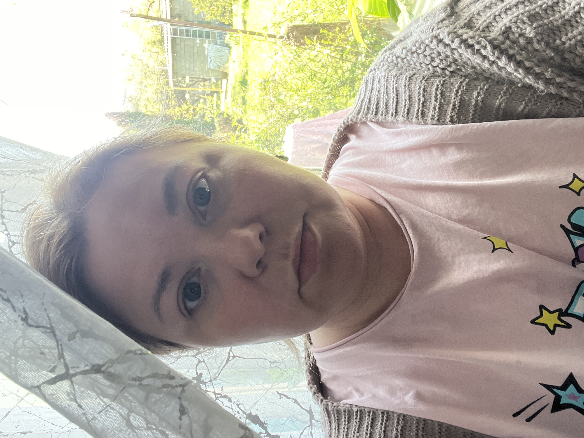 A selfie-style portrait captures a woman indoors, her head tilted, looking directly at the camera with a neutral expression. She has light-colored hair and blue-grey eyes. She is wearing a light pink t-shirt adorned with cartoon-like graphics, featuring yellow stars and other celestial motifs. Over the t-shirt, she has on a light brown or grey knitted cardigan with visible ribbed and cable patterns.
On the left side of the frame, a sheer white curtain with a subtle, intricate pattern is partially visible. Behind the woman, outside a window, a vibrant green outdoor scene is brightly lit by the sun, suggesting it is a sunny day. Lush green foliage, likely trees or bushes, fills the background. A glimpse of a building structure, possibly a shed or fence, can be seen through the greenery. The bright natural light from outside causes some areas to appear overexposed. The setting appears to be a residential area with natural surroundings, consistent with the location of Vesyoloye, Russia.
The primary activity depicted is the woman posing for a photograph. No readable text is visible, only the graphic elements on her t-shirt. The overall impression is one of calm and natural light.