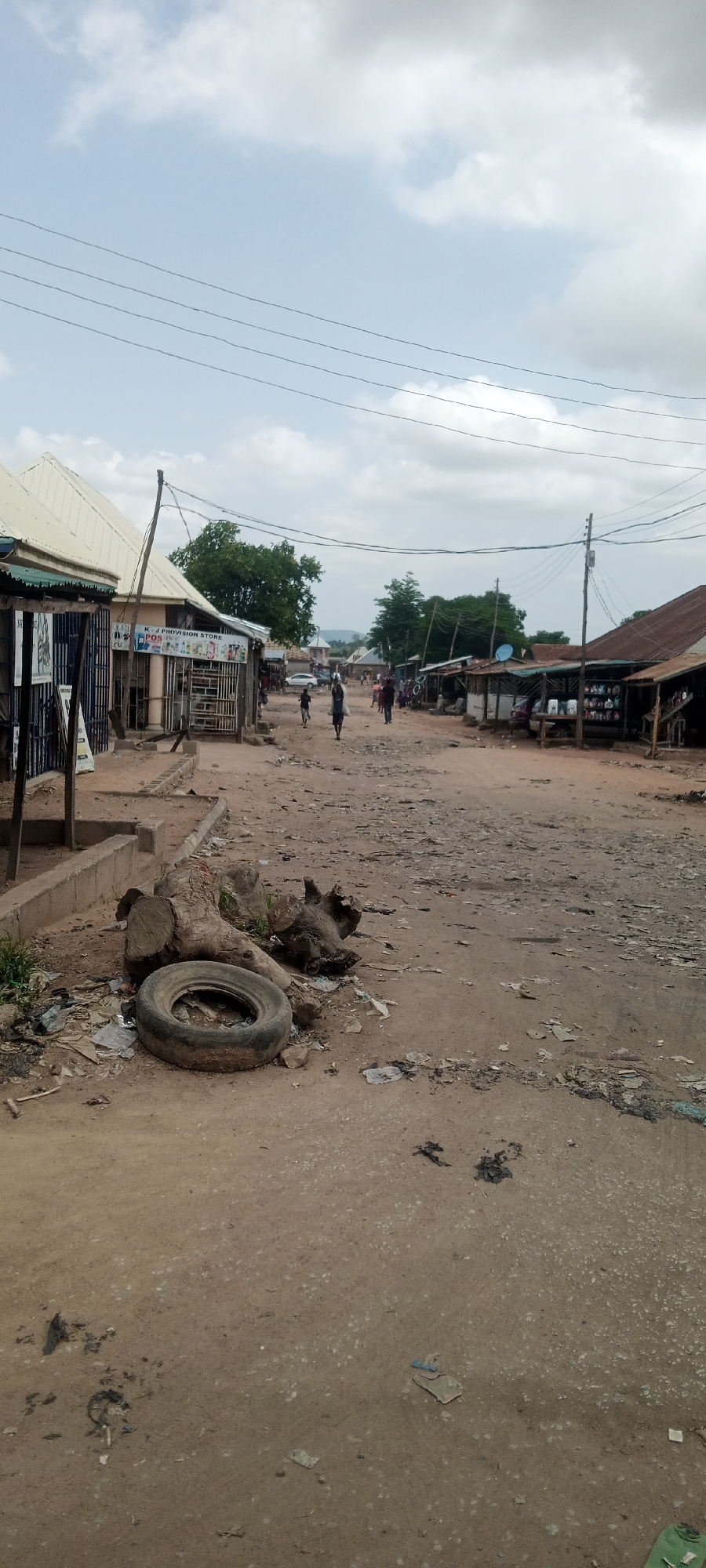 This is a ground-level, eye-level vertical photograph of a dusty street in Jalingo, Nigeria. The street is unpaved and appears to be in disrepair, with loose dirt and debris scattered across its surface. Several structures, likely shops or market stalls, line both sides of the street. These structures are simple, with corrugated metal roofs and open fronts displaying goods for sale. Some of the stalls have awnings extending over the sidewalk.
In the foreground on the left, a discarded tire and tree stumps are piled on the side of the road, surrounded by litter. Further down the street, several people are walking, some carrying bags or parcels. One person is walking towards the camera, carrying a large white bag over their shoulder. Another group of people are walking away from the camera in the distance. The sky is partly cloudy, with a few scattered white clouds against a pale blue background. Utility poles and wires are visible stretching across the upper portion of the image, spanning the width of the street. The overall impression is that of a busy, yet somewhat underdeveloped, commercial area.