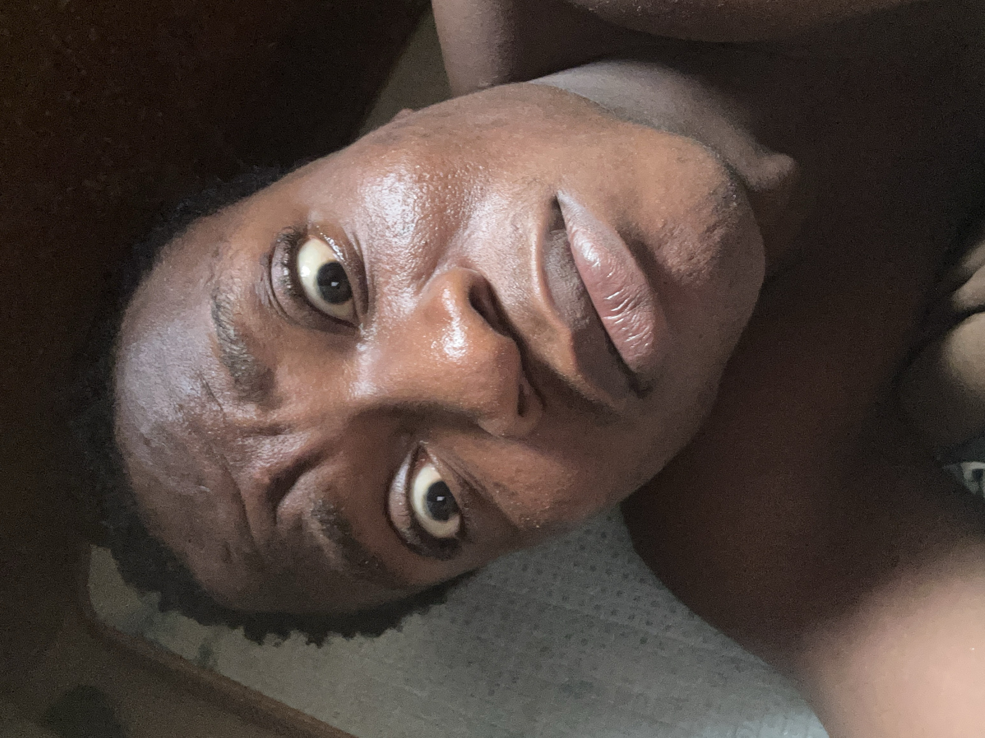A close-up, head-and-shoulders shot of a person with dark skin lying on what appears to be a light-colored surface. The person's face is slightly tilted, and their eyes are wide open and focused towards the camera. The skin appears to be slightly oily or sweaty. The background is blurry and indistinct, with hints of dark wooden surfaces. The location is unknown.
