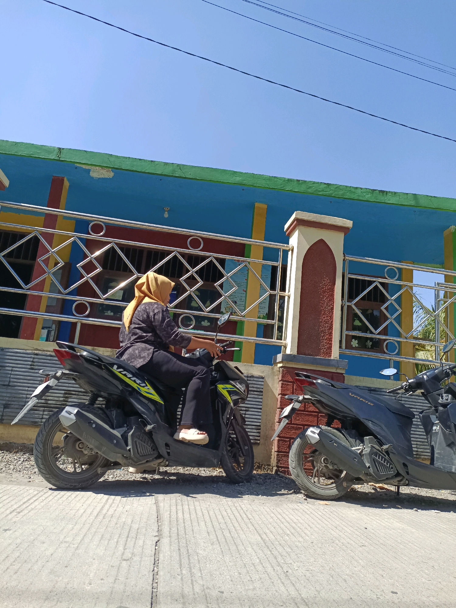 In Cangkring Rembang, Indonesia, on a bright, sunny day, a woman wearing a yellow hijab and a dark patterned shirt is seated on a black scooter. She is looking to her right, with her hands on the handlebars. The scooter is parked on a concrete surface in front of a building with a brightly colored facade. The building has a blue upper section with green trim and yellow and red vertical stripes. A metal railing with a geometric pattern runs along the front of the building, and a decorative pillar with a reddish-brown textured surface stands near the center of the frame. Another black scooter is parked to the right of the woman's scooter. The ground is a mix of gravel and concrete, with visible tire marks on the concrete. The sky is clear blue.