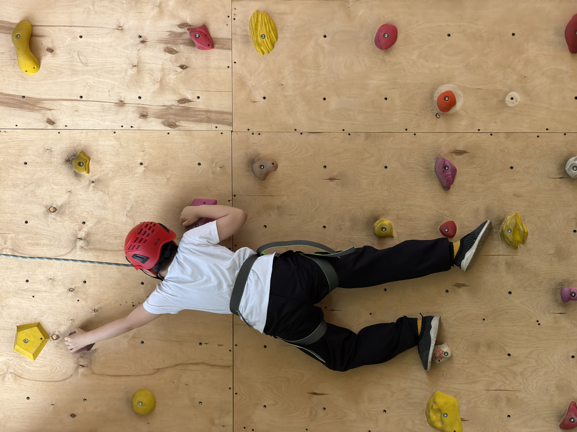 A child wearing a red helmet, a white t-shirt, black pants, and black and white sneakers is climbing an indoor rock climbing wall. The child is wearing a climbing harness and is secured to a rope. The climbing wall is made of plywood and has various colorful climbing holds attached to it. The wall appears to be made of sections attached together. The location is likely an indoor climbing gym, but the specific location in Izhevsk, Russia, cannot be determined from the image.
