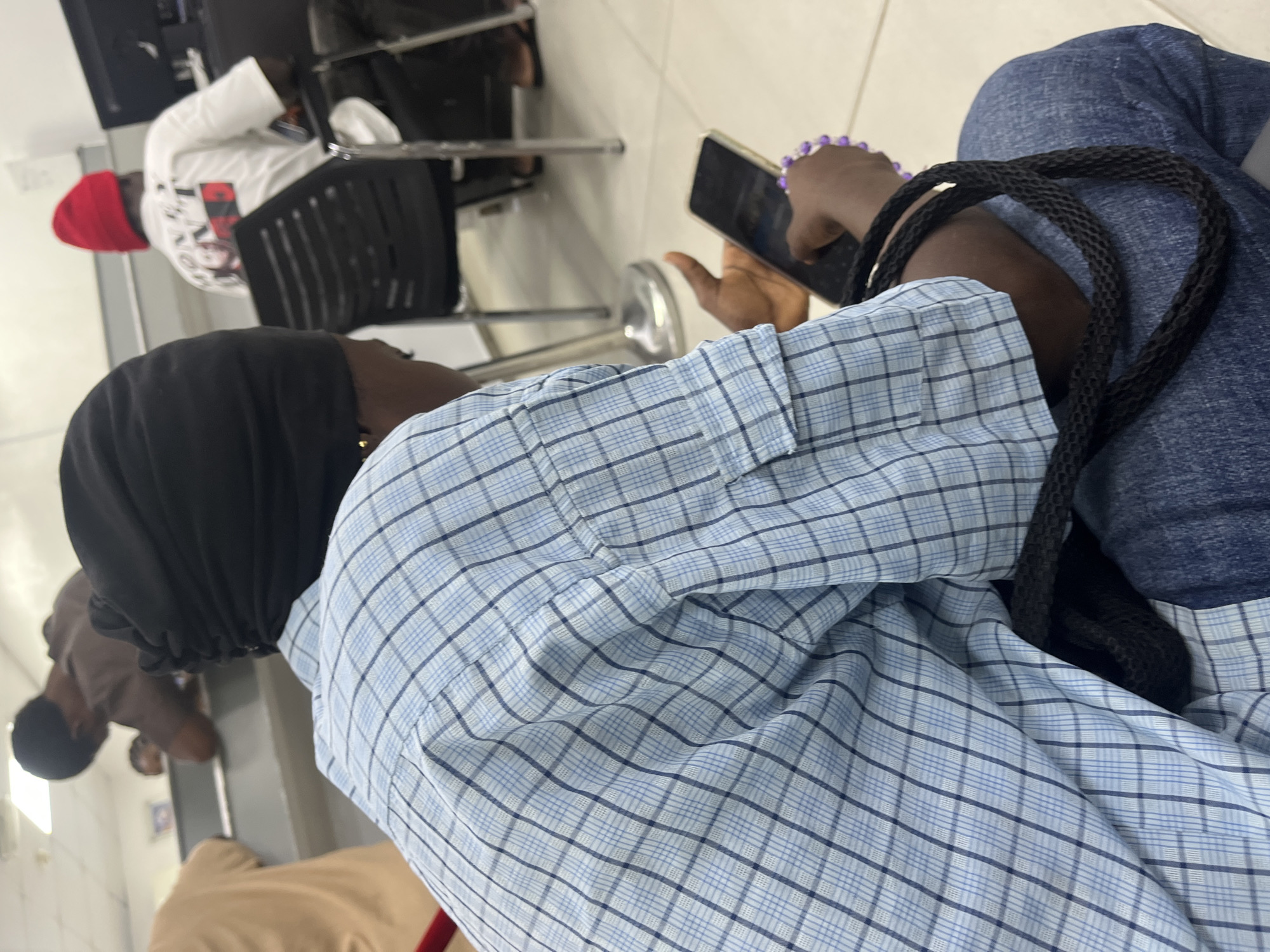 In an indoor setting, potentially in the city of Ado, Nigeria, a person wearing a light blue checkered shirt and blue jeans is sitting down while holding a smartphone. The person is wearing a black head covering. A black woven strap lies on their lap. In the background, there are other people seated, including a person in a white shirt and red cap seated in a black chair. The floor is light-colored tile.