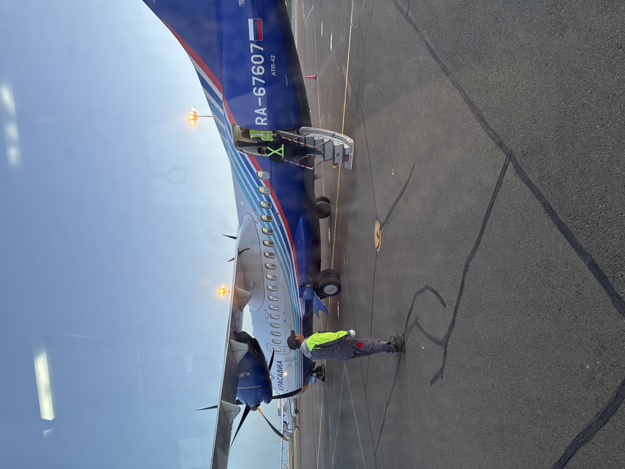 The image depicts an ATR-42 aircraft, registered as RA-67607, on an asphalt tarmac. A male individual wearing a gray jumpsuit and a neon yellow vest stands near the front of the plane. Another individual, wearing a similar vest, is visible on the aircraft steps. The plane is painted in blue, white, and red stripes, with the word "KPAСABИА" visible on the fuselage. The scene suggests an airport or airfield setting. The location in Zuunmod, Mongolia cannot be verified from the provided image.