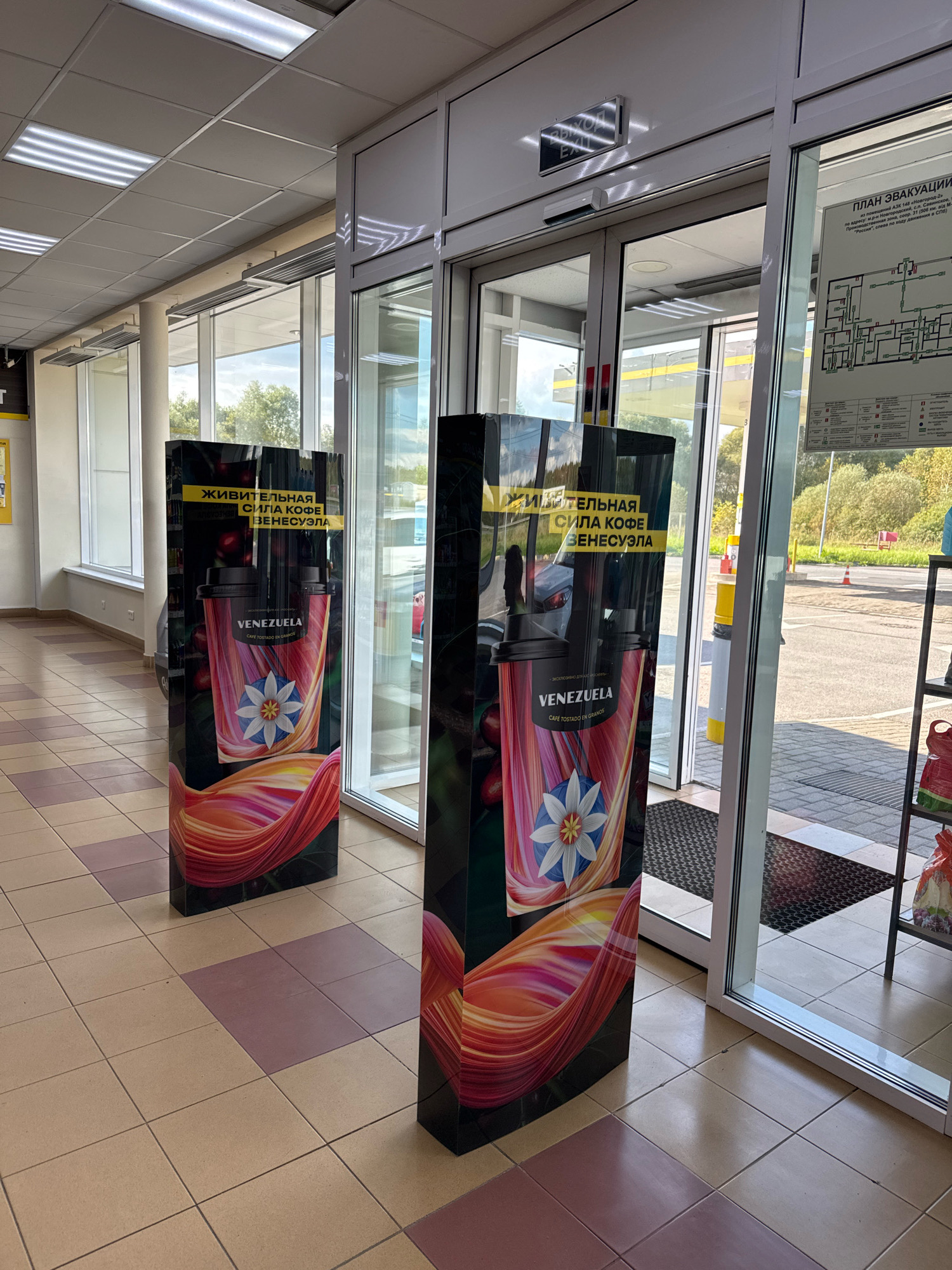 Two tall, rectangular promotional stands advertising "VENEZUELA" coffee are positioned inside a building, flanking an automatic glass door entrance. The stands feature a vibrant graphic of a coffee cup with a stylized flower design and flowing colors, along with Russian text. The floor is tiled with alternating light brown and muted pink squares. To the right of the stands, a large glass door opens to an exterior scene of a parking lot with trees and a road in the background. Above the door, a "PLAN EVAKUATSII" (evacuation plan) sign is visible. A black and silver "EXIT" sign is also mounted on the ceiling. The lighting is bright, coming from fluorescent ceiling panels.