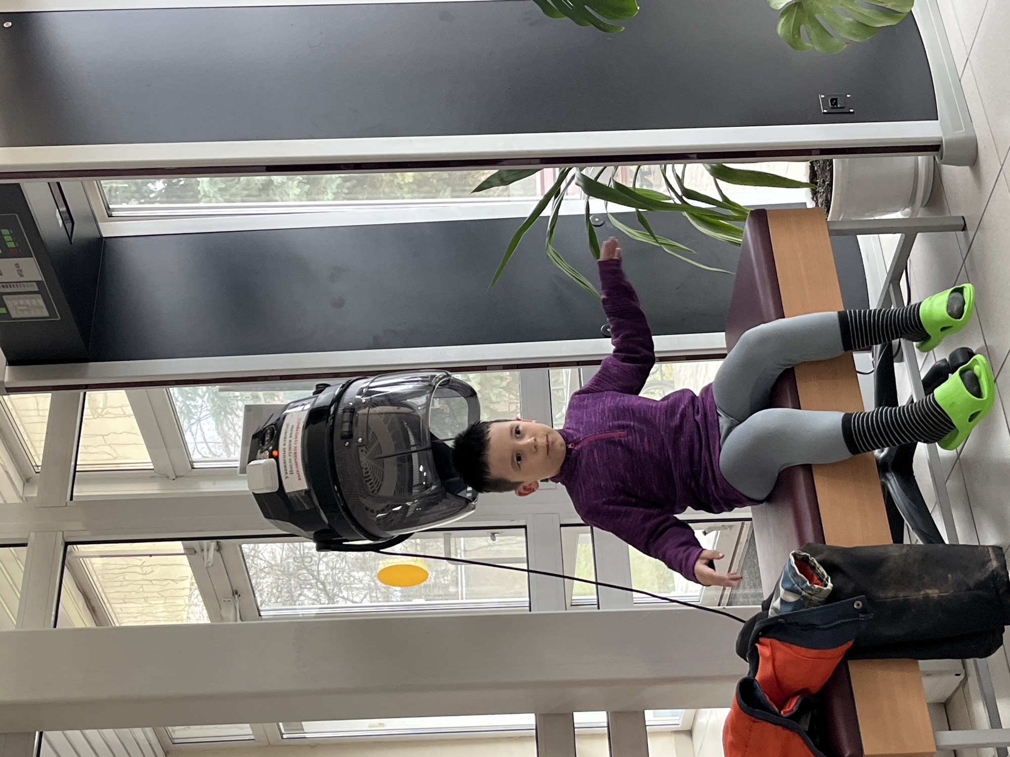 The image captures a young child, likely a boy, engaged in a physical therapy or exercise session indoors. The child is lying on their back on a low, padded bench, clad in a purple long-sleeved athletic top, light grey leggings, black and white striped socks, and bright neon green specialized footwear, possibly designed for therapy or rehabilitation. Their head is positioned beneath a large, clear-domed black machine, which has a fan-like structure visible inside. One of the child's arms is raised towards the top of the frame, while the other rests beside their body. They maintain a neutral to slightly curious expression, looking directly at the viewer.
On the white sticker attached to the machine, Cyrillic text is visible, which translates to: "DEAR CLIENTS! PERFORM TRAINING EXERCISES WITHOUT WEIGHT." This confirms the device's function as a therapy or exercise apparatus rather than a hair dryer.
The setting appears to be a facility with large windows, possibly a rehabilitation center or gym, in Chaikovsky, Russia. Through the windows, a bright, overcast daytime sky and bare tree branches are visible, suggesting either late autumn or winter. Potted plants, including a large Monstera, are placed near the windows. A black bag with an orange jacket is seen on the floor next to the bench. The room features a light-colored tiled floor and dark panels on the walls with white framing.