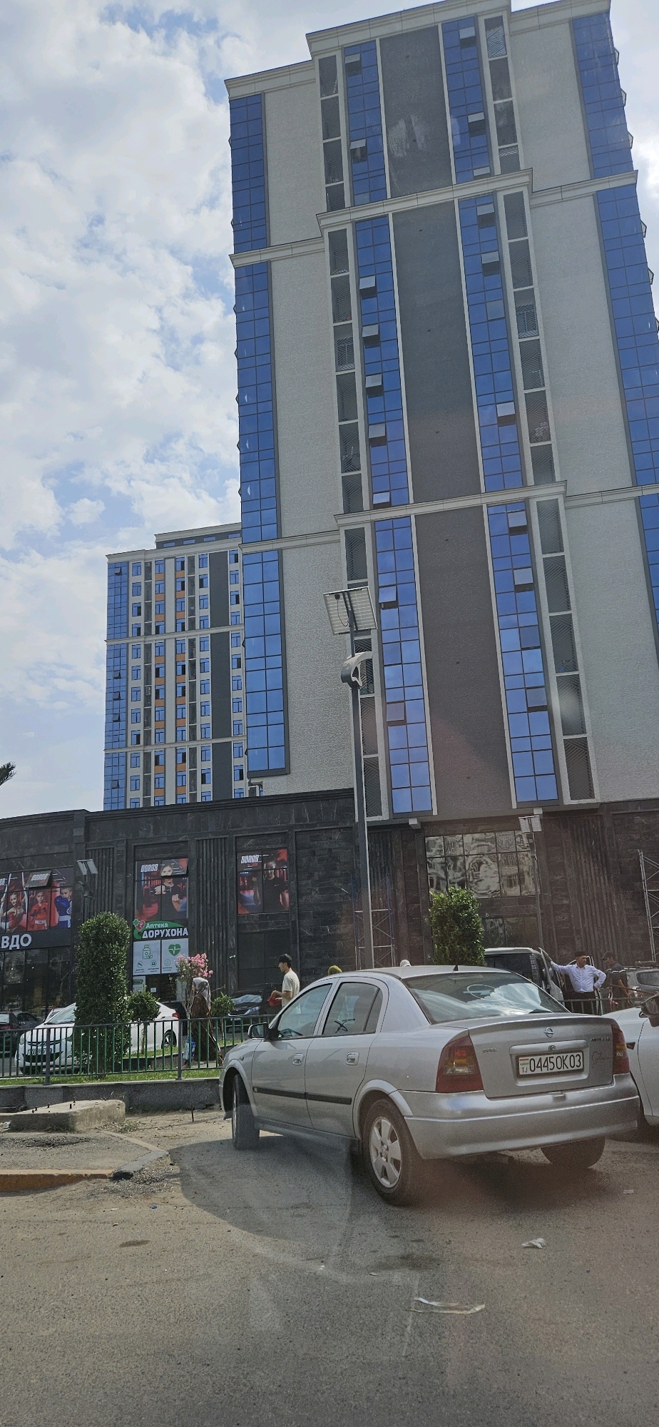The image captures a street scene in Dushanbe, Tajikistan, featuring modern architecture and everyday urban life. Dominating the background are two tall, contemporary buildings with grey facades and prominent blue glass windows. The building on the right is taller and features a darker grey vertical section. In front of these buildings, a single-story structure with a dark, textured facade houses various businesses, indicated by posters and signage. One poster displays "BORBA" in red lettering, suggesting a fitness or sports-related establishment. Another sign reads "АПТЕКА ДОРУХОНА," identifying a pharmacy.
In the foreground, a silver sedan, possibly an Opel Astra, is parked or driving slowly on a paved road. The car's license plate is visible and reads "TJ 0445OK03". Several individuals are present: a man in a white t-shirt and a black cap is walking near the businesses, and another man in a white shirt is standing by the silver car. A woman in a hijab is also visible. A green metal fence separates the sidewalk area from the street. The environment appears to be a commercial district with trees and some greenery interspersed between the buildings and the street. The sky is partly cloudy, indicating daytime.