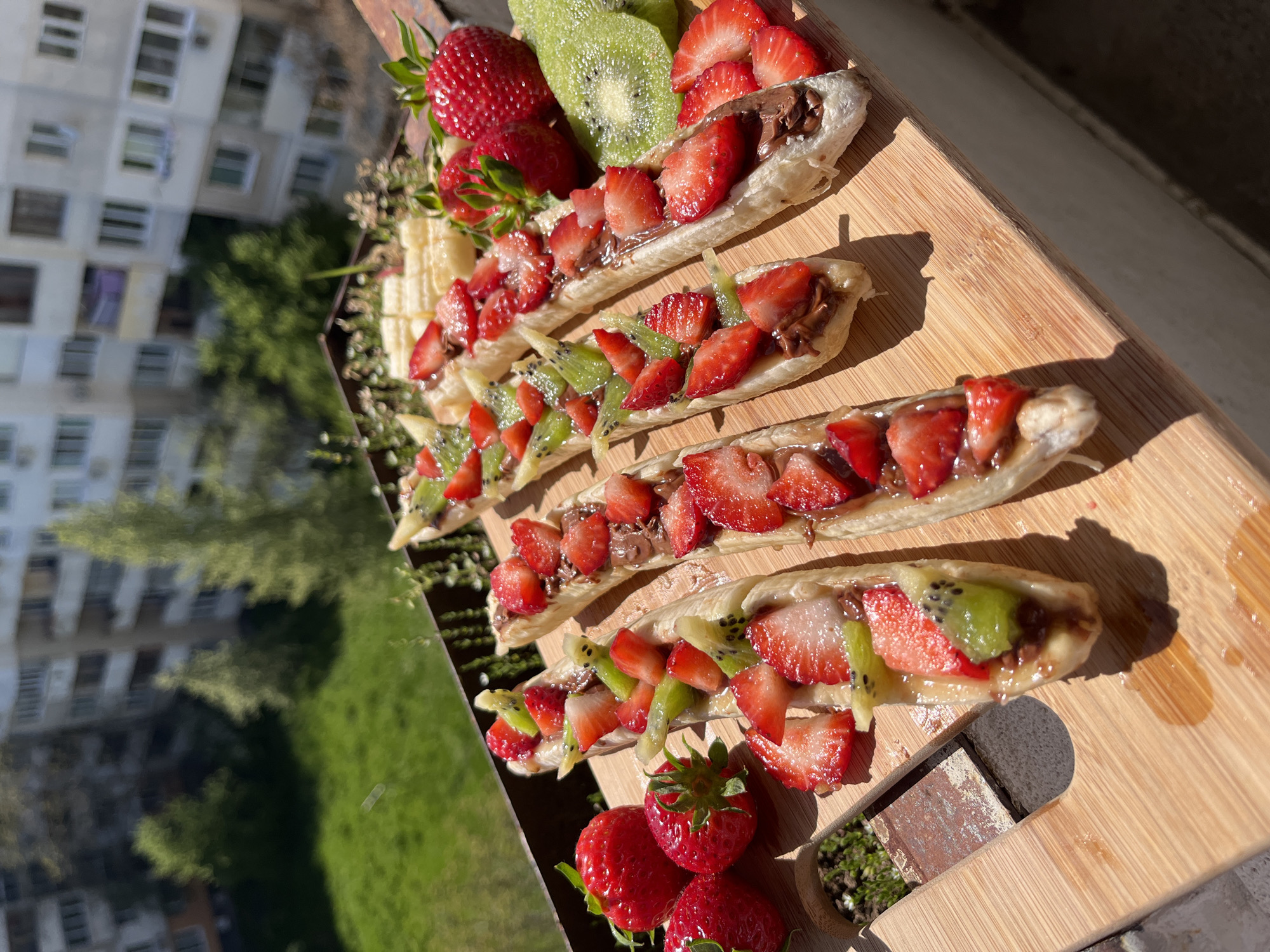 This image captures a vibrant display of fruit-topped pastries arranged on a light-colored wooden cutting board. The main subjects are five elongated pastries, each generously filled with a brown, chocolate-like spread and topped with a mixture of sliced fresh strawberries and sliced kiwi. Some pastries feature only strawberries, while others have both fruits.
Accompanying the pastries on the board are whole fresh strawberries, additional slices of kiwi, and a few pieces of sliced banana. A small, dark-colored decorative item, possibly a miniature planter with green moss inside, is also visible on the lower right corner of the board.
The setting appears to be outdoors or near a large window, with a multi-story apartment building visible in the background. This building is light-colored with numerous rectangular windows, suggesting an urban or suburban residential environment. Below the building, there is lush green grass and several trees, indicating a natural landscape within the urban context.
The strong, defined shadows cast by the food and the board on the wooden surface indicate bright sunlight, suggesting the photograph was taken during the day. The overall impression is one of a fresh, appealing snack or dessert, likely prepared for casual consumption in a pleasant, sunny setting. There are no people visible, and no discernible text is present in the image. Given the provided location context, the scene is likely set in Sofia, Bulgaria.