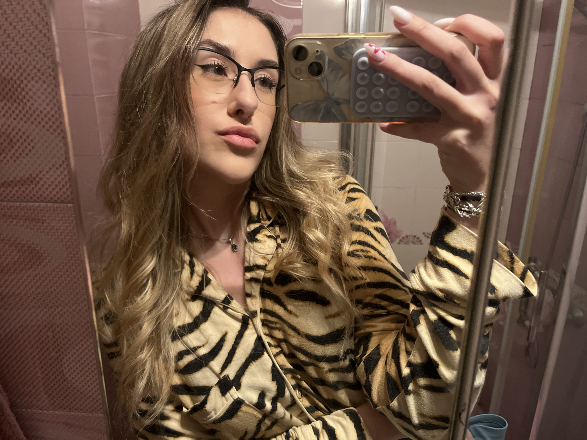 A woman takes a mirror selfie in a bathroom. The bathroom has pink tiles. The woman is wearing black-rimmed glasses and a tiger-striped shirt. She is holding a phone with a butterfly-patterned case and a pop socket. Her nails are long and white with a small red heart design on one nail. She is wearing a silver bracelet. The location is a bathroom, the location of which is not discernable from the image alone as being in Sofia, Bulgaria.