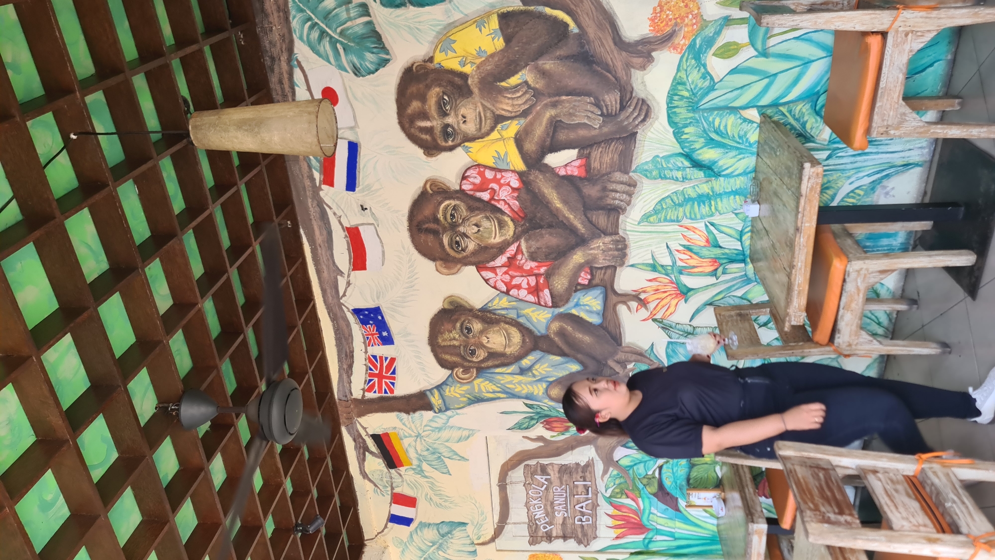 The image depicts the interior of a cafe or restaurant in Denpasar, Indonesia, with a prominent mural of three monkeys wearing shirts, seated on a branch amidst tropical foliage. The mural is decorated with various national flags suspended above the monkeys. A woman, wearing a black t-shirt and dark pants, stands in the foreground to the left, holding a glass with a beverage. She is positioned between two wooden tables with orange cushions on chairs. The ceiling features a wooden lattice structure with green painted panels, and a ceiling fan is visible. A decorative pendant lamp hangs from the ceiling. The cafe has a rustic ambiance with wooden furniture.
