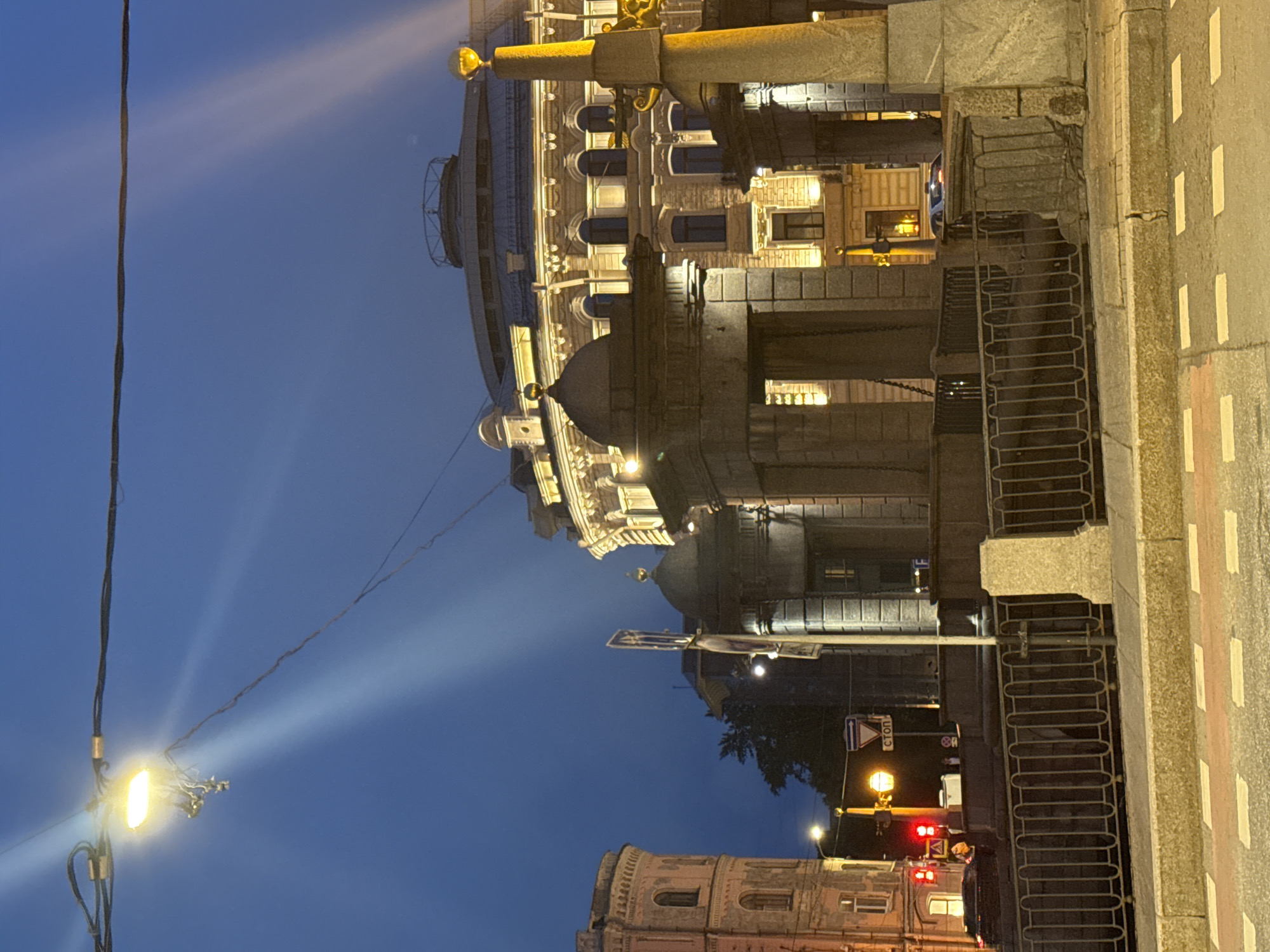 The image captures a night scene in Saint Petersburg, Russia. The foreground features a street with red and white markings. To the left of the street is a section of a bridge with an iron railing. Above the bridge are prominent buildings. The architecture is ornate, with multiple windows, columns, and decorative elements, lit by streetlights. In the background, there is a reddish building. A streetlight is visible, shining brightly, with electrical wires extending towards the upper left corner of the image. The sky is a dark blue color.
