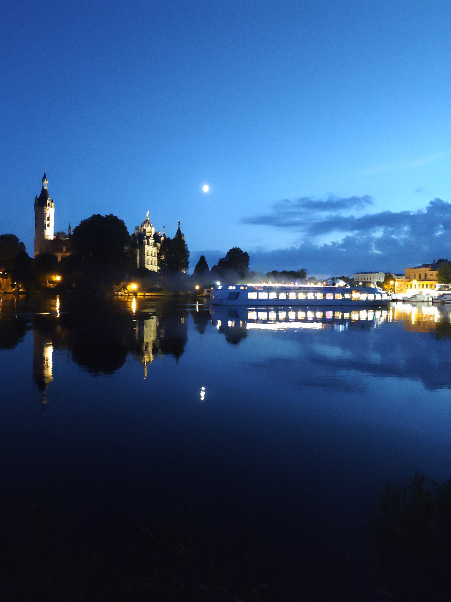 This image depicts a nighttime scene of the Schwerin Castle and its reflection in the waters of the Schweriner See in Schwerin, Germany. The sky is a deep blue, transitioning to a lighter blue near the horizon, with a few wispy clouds and a full moon visible. The castle, brightly illuminated, stands prominently on the left side of the frame, its towers and spires detailed against the dark sky. Its reflection is sharp and clear in the calm water below. A large passenger boat, also illuminated with many lit windows, sails across the water in the middle ground, its lights reflecting on the surface. On the right side of the image, more buildings are visible along the shoreline, also illuminated and reflecting in the water. The overall atmosphere is serene and picturesque, characteristic of a twilight or early night view of a historic waterfront. There are no people clearly visible in the image.