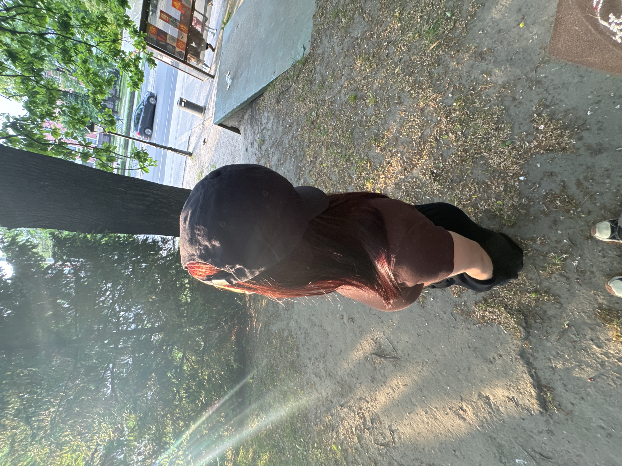A high-angle shot shows a person wearing a dark baseball cap and a brown shirt standing in a park-like area in an urban setting. The ground is a mix of dirt, sand, and scattered debris like leaves and twigs. A tree trunk is visible on the left side of the frame, with lush green trees and foliage extending upwards. In the upper left corner, a street scene is partially visible, including a parked car and what seems to be the edge of a building or sidewalk. In the upper part of the frame, there is also a concrete wall. The overall environment suggests a park or green space situated near a city street. Parts of another person with brown pants and shoes can be seen on the upper right side of the image.