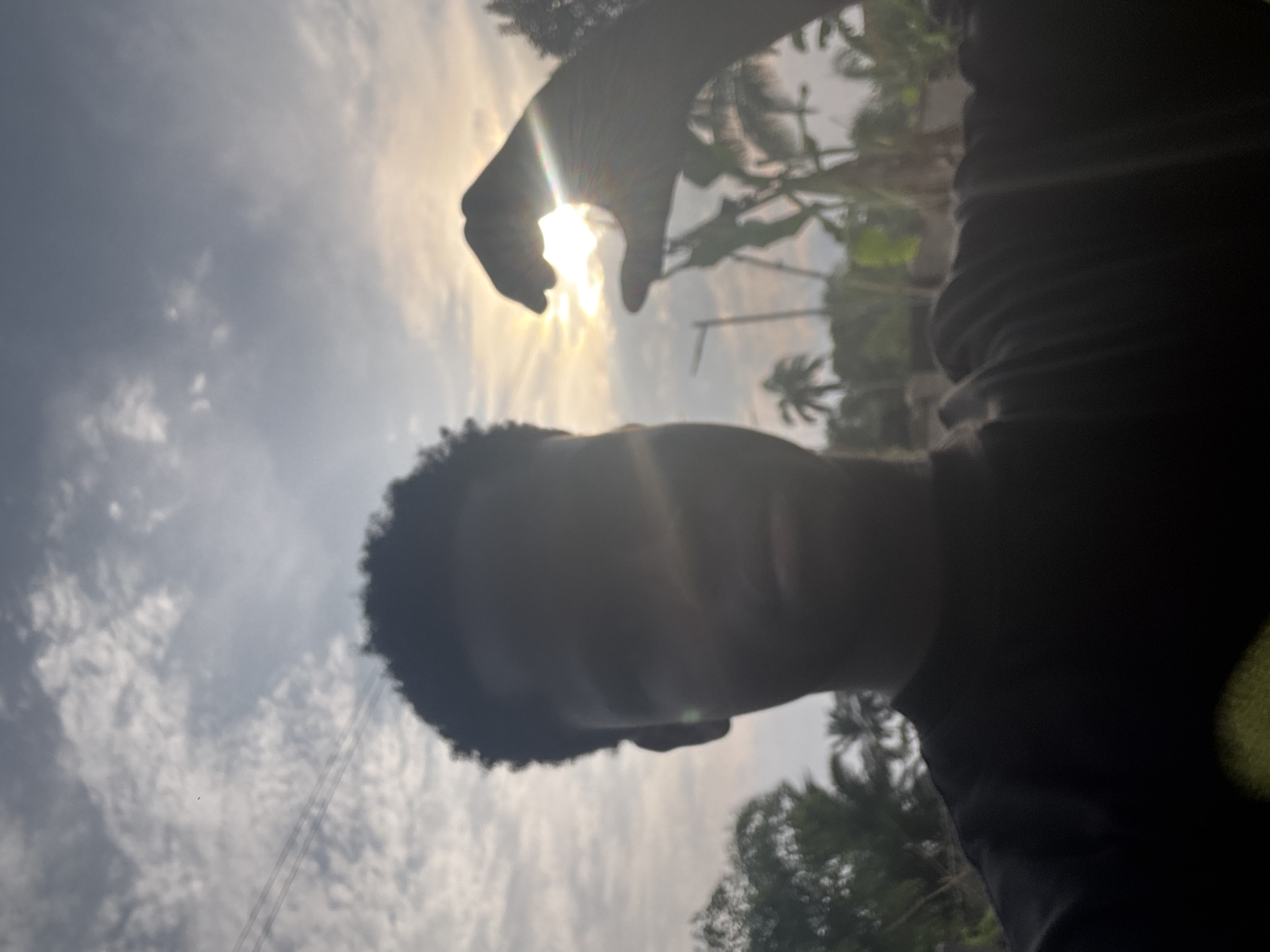 The image shows a person's silhouette against a bright sky in Aboh, Nigeria. The person is holding their hand up, partially obscuring the sun. Palm trees are visible in the background, indicating a tropical environment. The sky is filled with clouds.