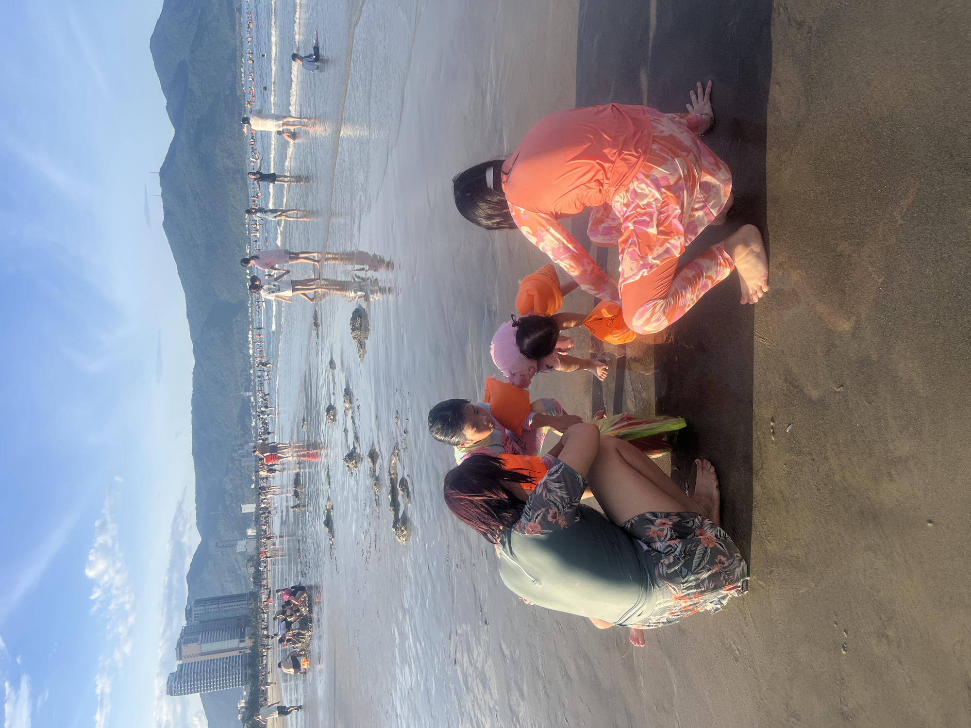 This is a horizontal, eye-level, medium shot of a family playing on the beach in Đà Nẵng, Vietnam. In the foreground, four people are crouched on the wet sand near the water's edge. Two adults and two children appear to be engaged in playing with sand and water. One adult, wearing a dark green rash guard with a floral pattern and dark shorts, is on the left. To their right, a child in a light blue, pink, and yellow patterned rash guard and orange arm floaties is interacting with a younger child. The younger child is wearing a pink swim cap and orange arm floaties. To the right of them, an adult in a bright coral-colored rash guard with a pink and orange abstract pattern is also crouching, facing away from the camera. The background shows a crowded beach with many people walking, playing, and swimming in the ocean. Buildings, including tall high-rises, are visible on the left side of the image, and a mountain range with lush green vegetation forms the backdrop. The sky is bright blue with some scattered clouds, and the sun is casting long shadows on the wet sand, indicating it is likely late afternoon or early evening. The ocean is calm with gentle waves.