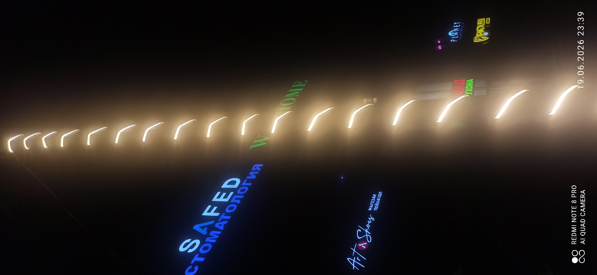 In an exterior night shot, a tall building is illuminated by a vertical series of light fixtures that emit a warm, golden glow. The lights are arranged in an ascending pattern, creating a visually striking effect against the dark sky. On the left side of the building, neon signs are visible. One sign, in blue, reads "SAFED СТОМАТОЛОГИЯ" (SAFED Dentistry). Below it, another sign in white script says "Art Shoes" with smaller text below it in Cyrillic, "МАГОЗАИ ПОЙАФЗОЛ" (Shoe Store). Further to the right, a green sign reads "WHOME". In the upper right quadrant, a sign displays the word "PLANET" in blue. Below the "PLANET" sign, a yellow and black sign is partially visible. The bottom of the image shows the timestamp "19.06.2026 23:39" and "REDMI NOTE 8 PRO AI QUAD CAMERA". The overall impression is of a commercial building facade at night, adorned with various illuminated signage.