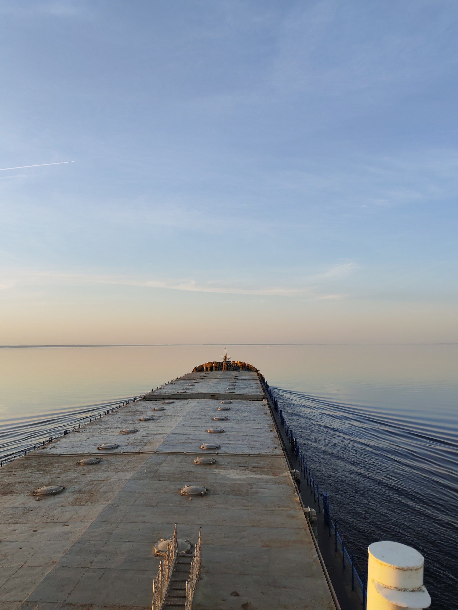 The image depicts a long, flat deck with numerous circular hatches, extending into the distance. The deck is likely part of a ship or barge on a calm body of water, as indicated by the wake trailing behind. The water is still and reflects the sky, which is a gradient of light blue at the top transitioning to a light orange/yellow hue near the horizon. A faint contrail is visible in the upper left corner of the sky. The image does not provide sufficient information to identify a city or specific location. No people, subjects, objects, actions, or interactions are visible in this environment.