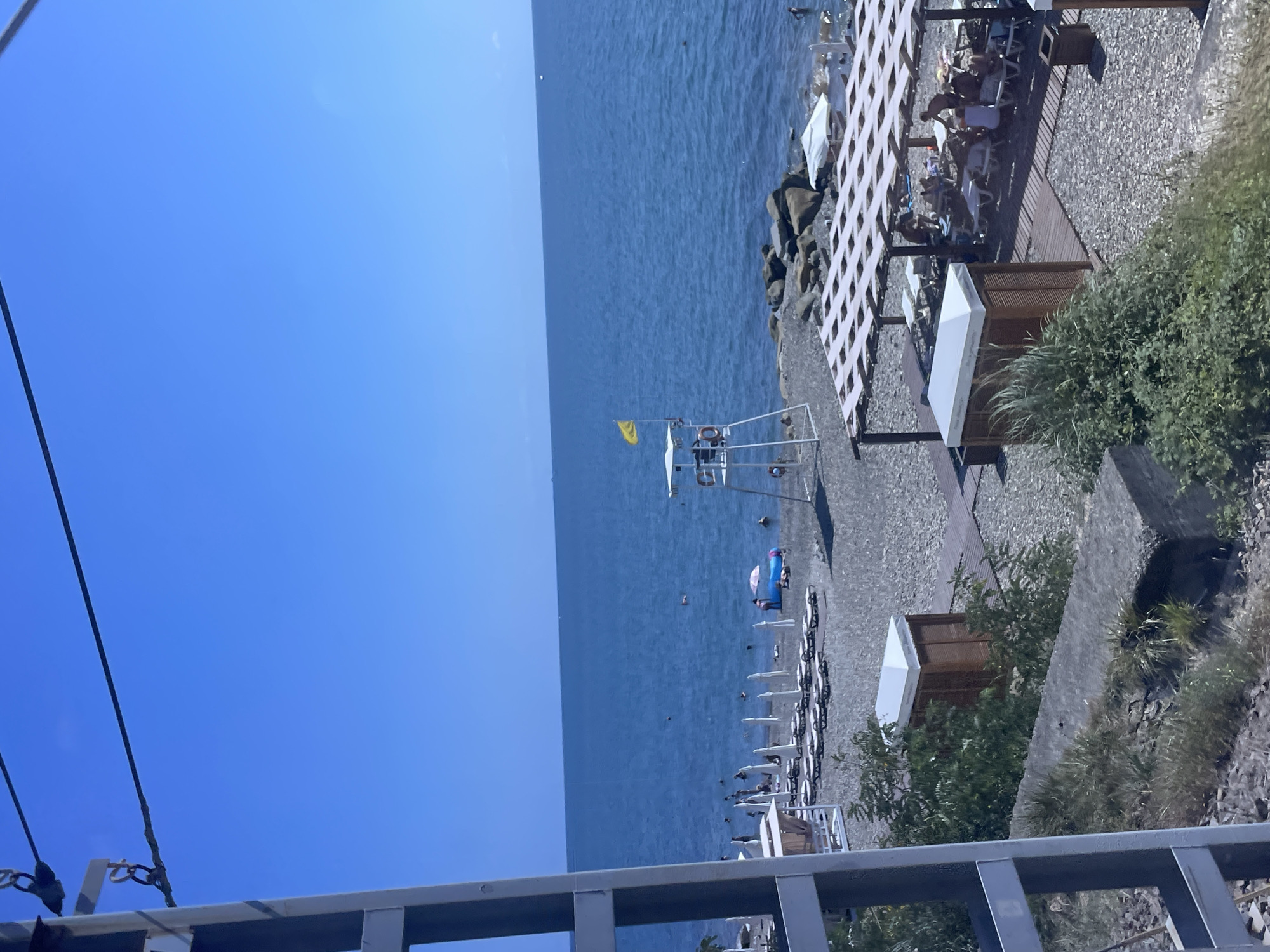The image depicts a sunny day at a pebble beach on the Black Sea coast in Sochi, Russia. The sky is a clear, deep blue, and the sea is a vibrant azure. In the foreground, a metal structure, possibly part of a viewing platform or infrastructure, partially obstructs the view.
The beach is dotted with rows of white sun loungers and umbrellas, indicating a popular tourist destination. Several people are visible, some lounging on the beach, others wading in the shallow water, and a few swimming further out. There is a lifeguard tower with a yellow flag flying from it, positioned near the water's edge. A blue inflatable raft is also visible near the shore, with a person or two on it.
Further back from the beach, there are wooden structures providing shade for sunbathers, with more loungers beneath them. Some of these structures have white canopies. The beach itself is covered in small, grey pebbles. Beyond the beach, a rocky area juts out into the sea. In the very background, a faint horizon line separates the sea from the sky, and a few distant objects, possibly boats, are visible on the water.
The overall scene is one of leisure and relaxation, typical of a seaside resort during peak season.