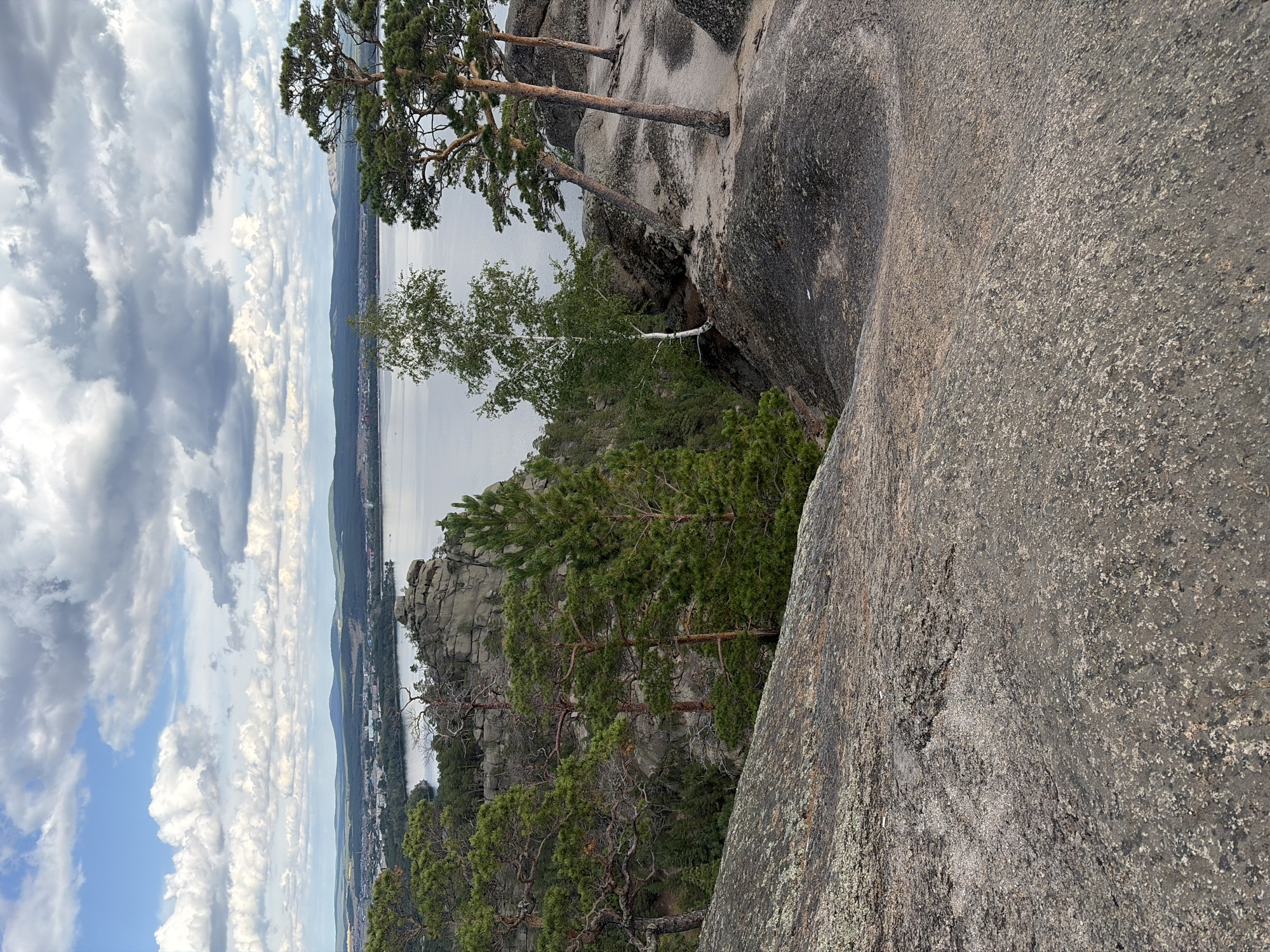 This is a scenic, eye-level, vertical, outdoor shot of a landscape from a high vantage point. The foreground is dominated by a large, textured rock formation with a rough, granular surface. Beyond this, a steep, rocky slope leads down to a body of water, which appears to be a lake. The lake stretches into the distance, with a town or settlement visible along its shore. A line of low, rolling hills with dark green forests forms the horizon, under a sky filled with large, white, puffy clouds against a pale blue backdrop. Several pine trees and a slender birch tree are scattered across the rocky terrain, with some of the pines growing at an angle from the rocks. The overall impression is one of natural beauty and vastness, suggesting a location in a park or natural reserve. The location is likely within the Зелёный Бор ауылдық округі in Kazakhstan, known for its picturesque natural landscapes. There are no people or animals present in the image.