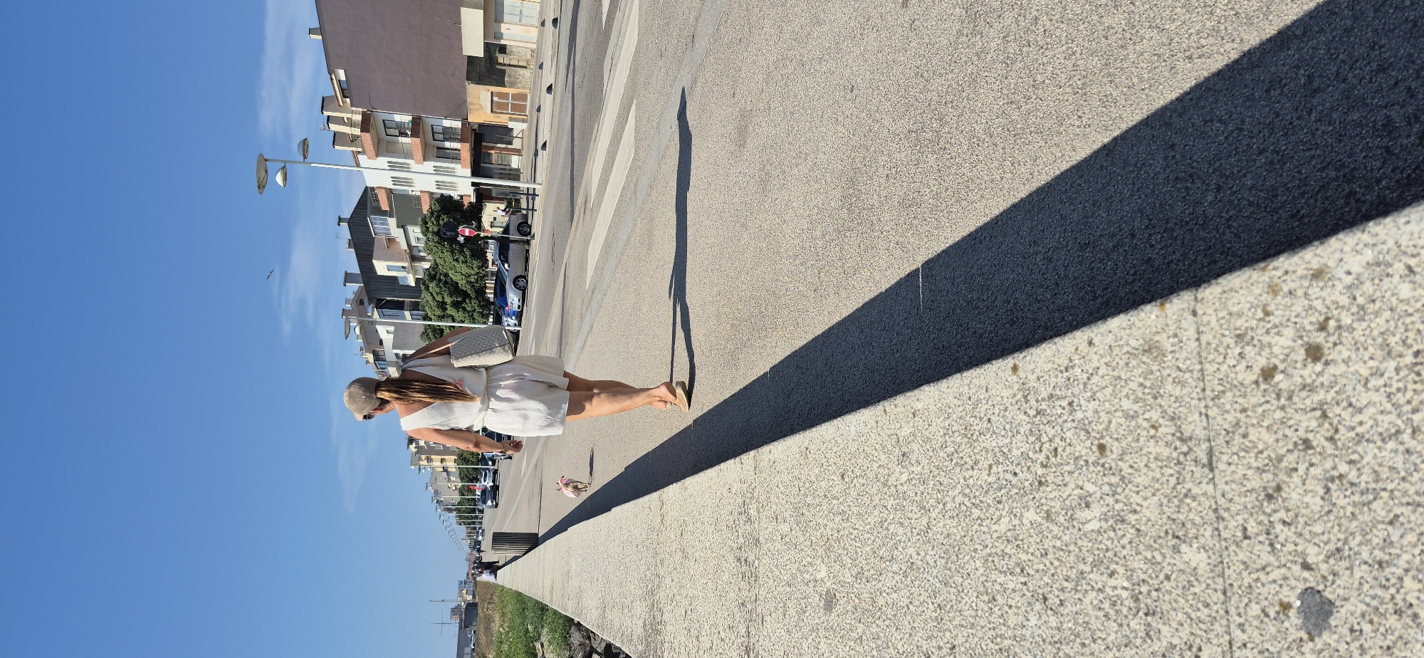 A woman, from behind, is walking along a paved promenade in Vila do Conde, Portugal, on a sunny day. She is wearing a light-colored romper, a cap, and sandals. She carries a large tote bag over her shoulder. A small dog, wearing a pink accessory, walks a short distance in front of her on a leash. The promenade runs parallel to a road with parked cars and some pedestrian traffic. Across the road, buildings with balconies and windows are visible, suggesting an urban or semi-urban coastal setting. The sky is clear and blue. The sun casts a long shadow of the woman and her dog across the promenade.
