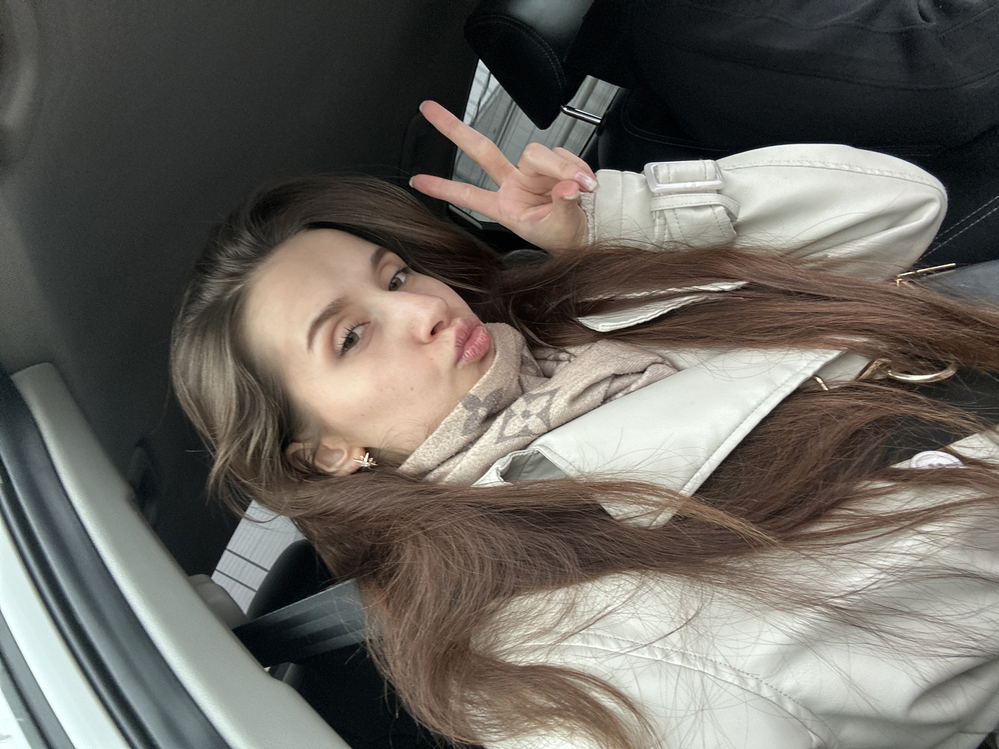 A young woman with long brown hair is making a peace sign while seated in a vehicle. She is wearing a light-colored jacket and a patterned scarf. The interior of the car features black seats and a grey door panel. The photograph appears to have been taken at an angle from above. There is no discernible information indicating that this photo was taken in Chaikovsky, Russia.