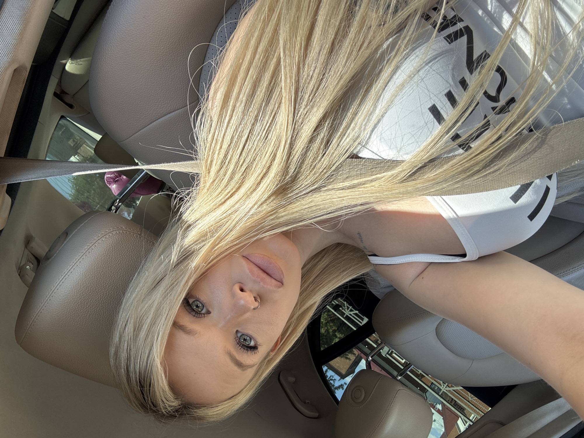 A blonde woman is taking a selfie inside a car. She is positioned in the driver's seat, leaning back. Her face is turned towards the camera, with her eyes looking directly at it. She has light-colored skin, light blue eyes, and her lips are slightly parted. She is wearing a white tank top with a black graphic design on it, and a nose ring. Her long blonde hair is flowing around her face and over her shoulder. The interior of the car is visible, with tan leather seats and a seatbelt. Sunlight is streaming into the car, creating shadows and highlights. Through the car windows, buildings and trees are visible, suggesting an urban or suburban setting. The overall impression is a casual, everyday moment captured by the person themselves. The image appears to be taken during the daytime, possibly in the afternoon, given the strong sunlight. There is a faint tattoo visible on her upper arm. The graphic on her shirt features bold black lettering, though the full text is not clearly legible.